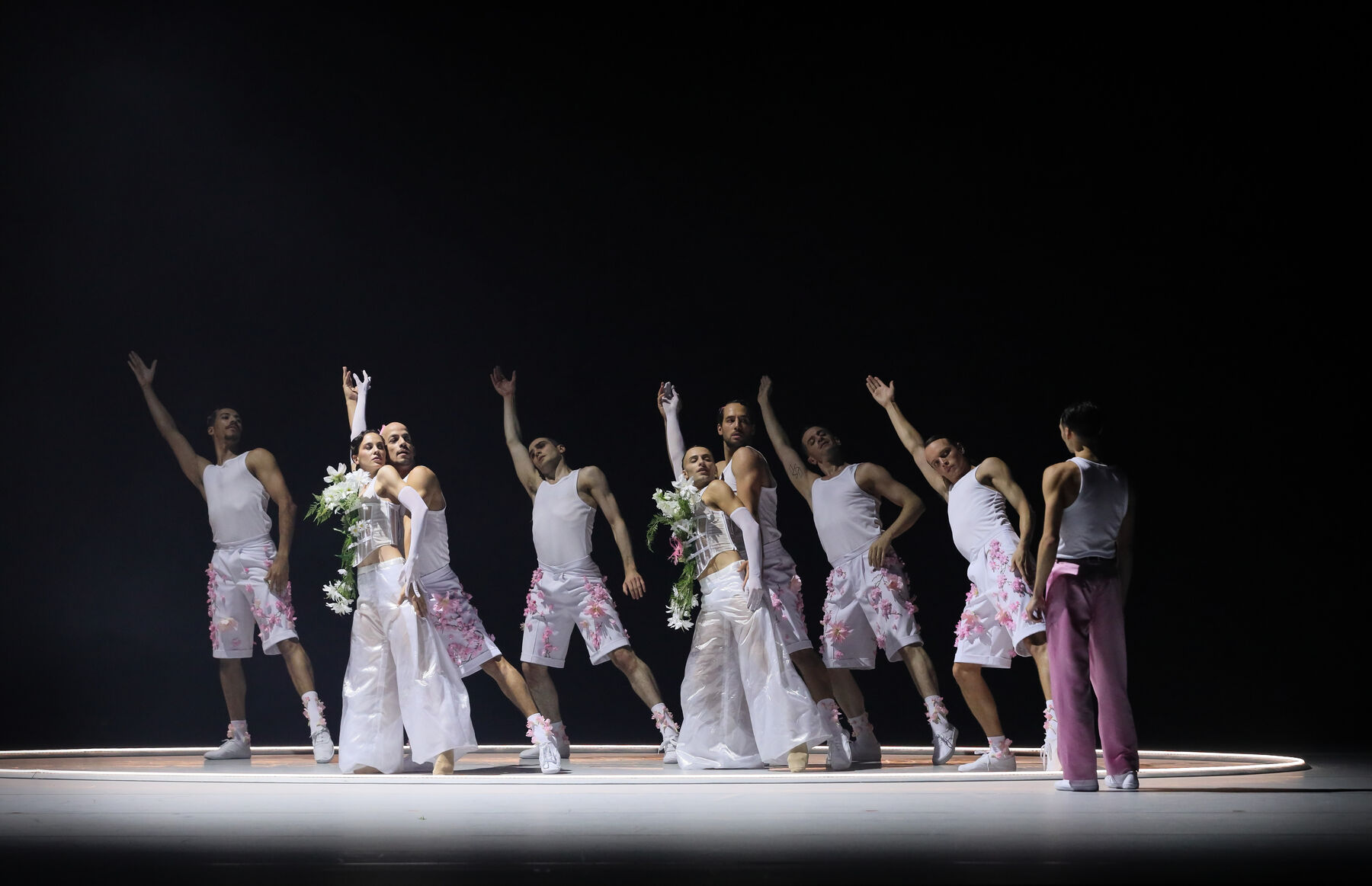 Tänzerinnen und Tänzer stehen in drei Reihen hintereinander. Sie neigen sich alle nach rechts und heben ihren rechten Arm gestreckt nach oben. Sie tragen weiße Kostüme mit verschiedenen aufgesetzten weißen und rosa Blumen. Alle stehen in einem weißen Kreis auf dem Boden, nur ein Tänzer in rosa Hose steht außerhalb des Kreises und schaut die anderen an. Der Rest der Bühne ist dunkel.
