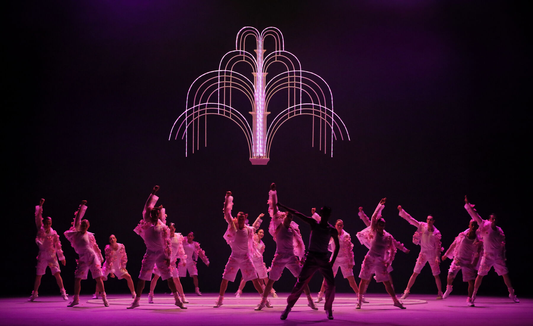 Viele Tänzerinnen und Tänzer in hellen, mit rosa Blüten verzierten Kostümen stehen gleichmäßig verteilt auf der Bühne. Sie haben ihre Beine mehr als schulterbreit geöffnet und heben ihre rechte Faust. Über ihnen hängt eine leuchtende zweidimensionale Installation, die einem Springbrunnen ähnelt. Die Szene ist in pinkes Licht getaucht.