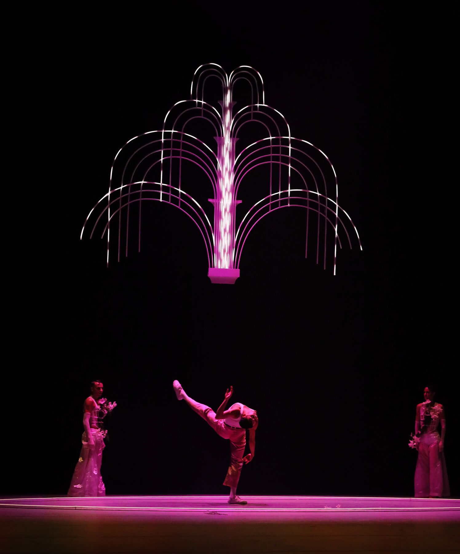 Auf dem Boden ist ein pink beleuchteter Kreis, in diesem Kreis tanz eine Mann. Hinter ihm, außerhalb des Kreises stehen zwei weitere Tänzer und beobachten ihn. Darüber hängt eine beleuchtete zweidimensionale Installation, die einem Springbrunnen ähnelt. 