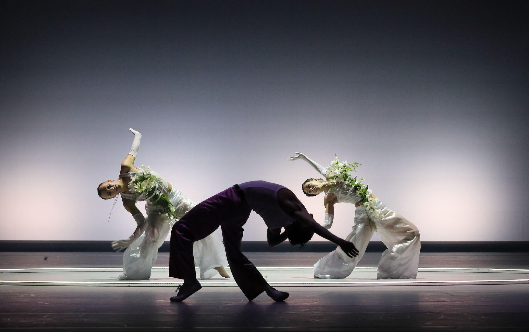 Auf dem Boden ist ein großer weißer Kreis. In diesem Kreis befinden sich ein Tänzer und eine Tänzerin in weißer, mit Blumen verzierter Kleidung. Weißes Licht ist auf sie gerichtet. Vor dem Kreis tanz ein weiterer Tänzer, er wird nicht angeleuchtet.