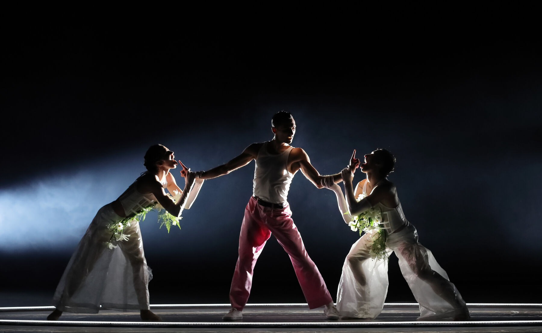 Ein Mann mit rosa Hose und weißem Oberteil steht breitbeinig zwischen zwei Mitgliedern des Balletts, welche weiße, mit Blumen dekorierte Kleidung tragen. Der Tänzer in rosa Hose streckt seine Arme nach links und rechts aus, die beiden in weißer Kleidung greifen seine Handgelenke und deuten an, seine ausgestreckten Zeigefinger abzulecken. Die drei werden gezielt von der Seite beleuchtet.