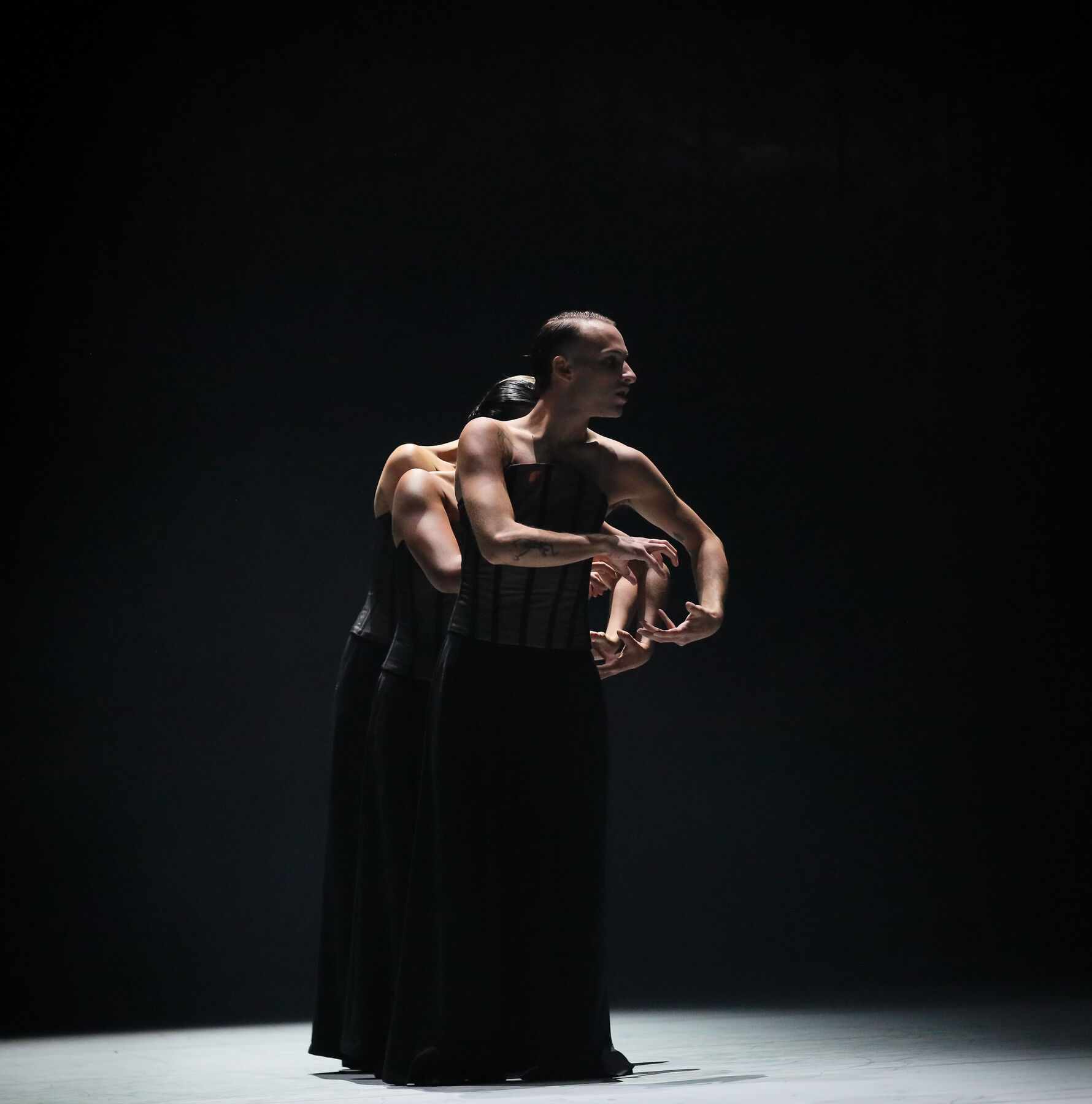 Drei Mitglieder des Balletts stehen hintereinander im zielgerichteten Scheinwerferlicht. Es ist nur das Gesicht des vordersten Tänzers zu sehen. Alle drei tragen ein reripptes semitransparentes Korsett und einen bodenlangen schwarzen Rock. Der Rest der Bühne ist dunkel. 