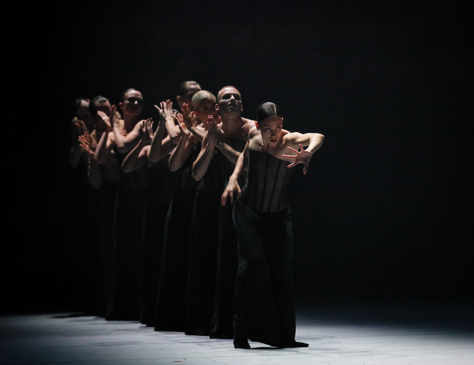 Einige Tänzerinnen und Tänzer stehen hintereinander aufgereiht. Sie tragen schwarze Korsett-Oberteile und schwarze, bodenlange, weite Hosen. Die Tänzerin an vorderster Stelle greift nach vorne. Die anderen falten ihre Hände in verschiedene Richtungen. Alle sind nur spärlich beleuchtet, der Rest der Bühne ist dunkel.