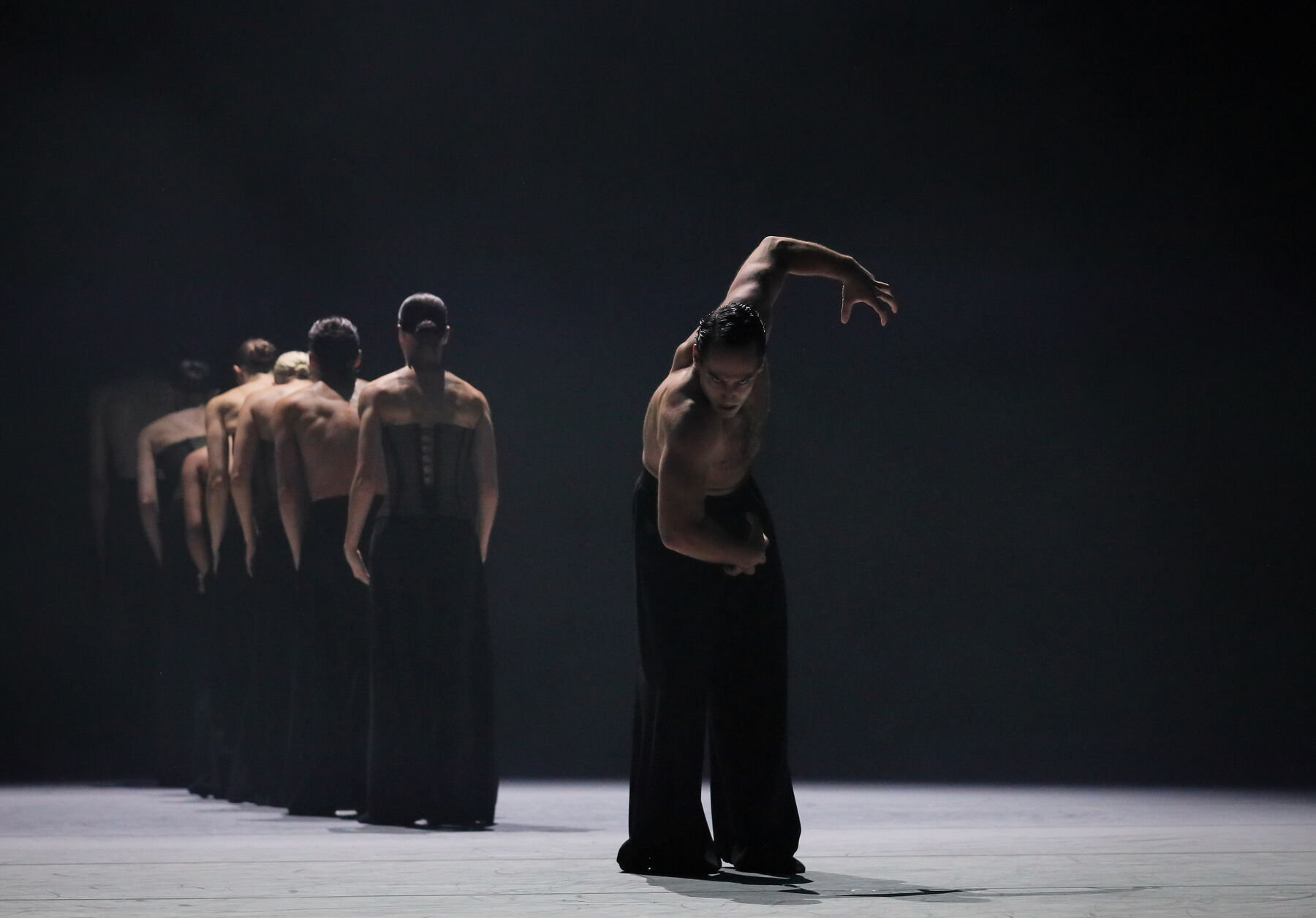 Eine Gruppe Tänzerinnen und Tänzer auf einer dunklen Bühne. Die Tänzerinnen tragen ein schwarzes Korsett und weite schwarze Hosen, die Tänzer sind oberkörperfrei. Alle bis auf ein Tänzer stehen in einer Reihe mit dem Rücken zum Publikum. Ein Tänzer beugt sich mit durchdringendem Blick nach vorne, er beugt seine Arme ausgestreckt und spannt seine Hände an, sodass sie wie Klauen anmuten. 