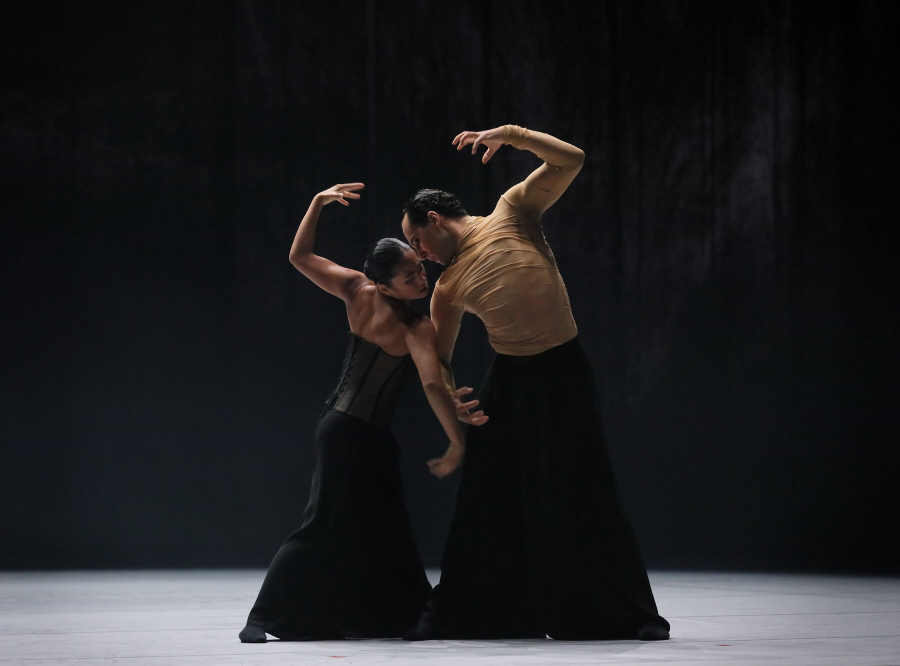 Ein Tänzer in einem hautfarbenen Oberteil und einer langen, schwarzen, sehr weiten Hose und eine Tänzerin mit schwarzem Korsett und ähnlicher Hose drehen sich Schulter an Schulter miteinander. Ihre Körper sind leicht zueinander geneigt, den abgewandten Arm heben sie angewinkelt nach oben.