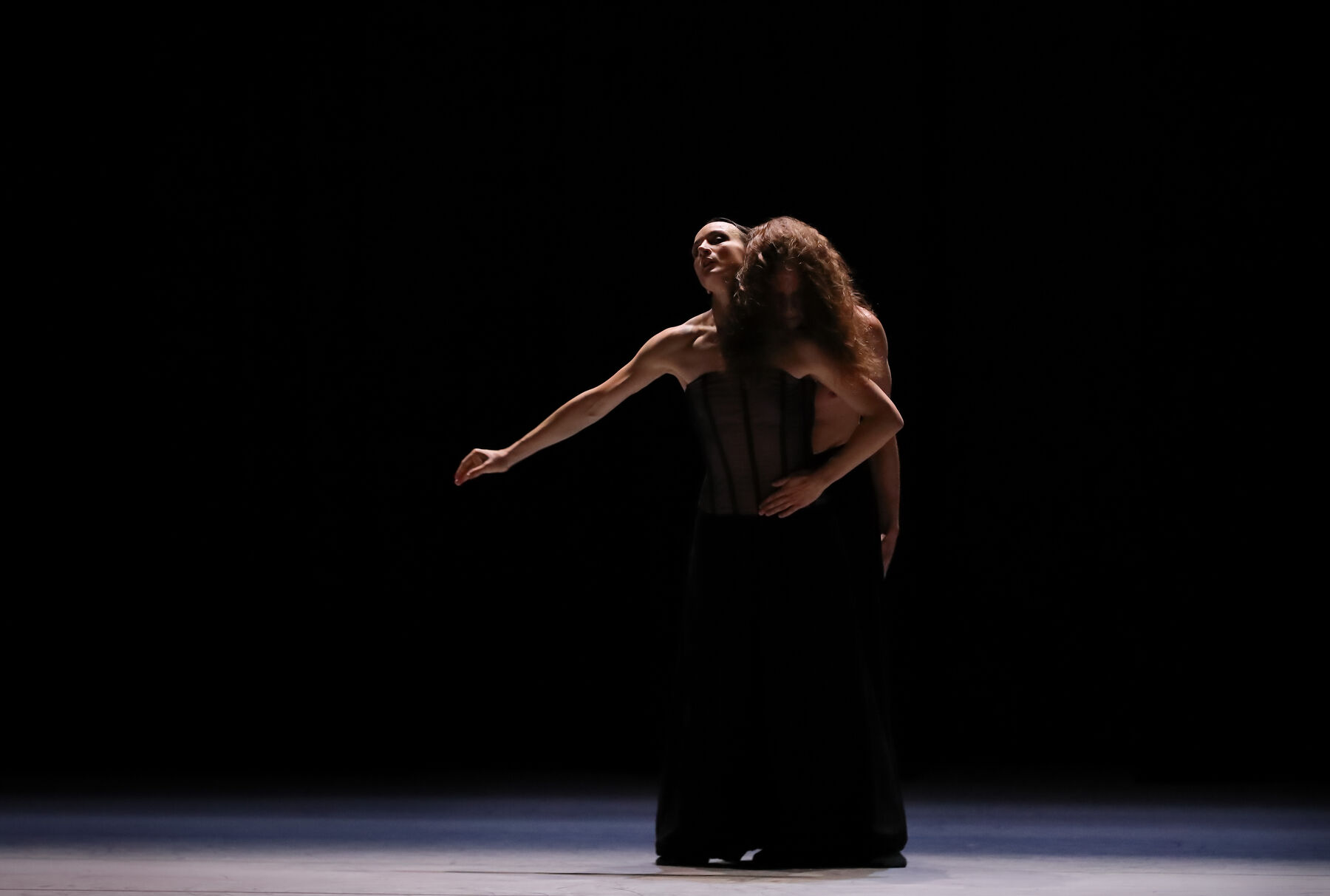 Eine Tänzerin und ein Tänzer stehen auf einer dunklen Bühne, das Licht ist nur auf sie gerichtet. Die Frau trägt ein schwarzes Korsett und eine weite schwarze Hose. Er trägt nur eine solche Hose. Sie legt eine Hand seitlich auf ihren Bauch, der Arm ist weggestreckt und gebeugt. Ihr anderer Arm ist gerade weggestreckt. Der Mann mit langem, gewellten Haar legt von Hinten seinen Kopf auf ihre Schulter. Sein Gesicht ist größtenteils von seinem Haar verdeckt.