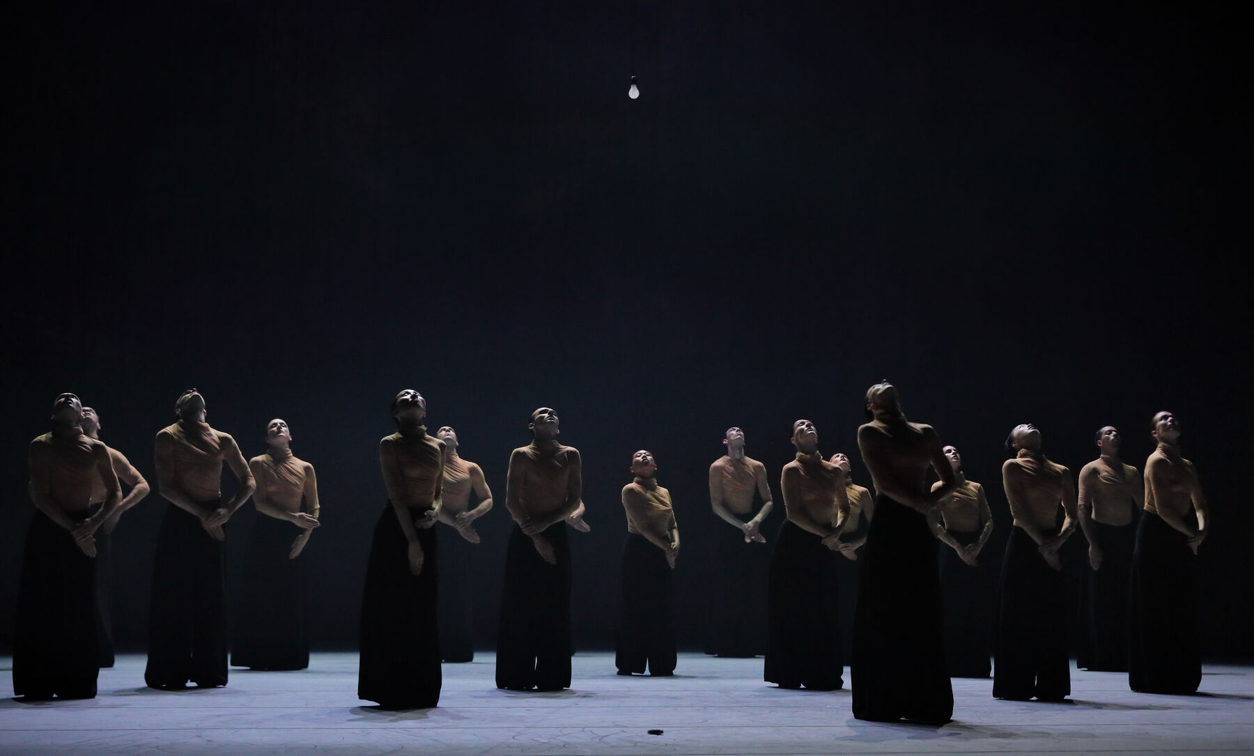 Viele Tänzerinnen und Tänzer stehen gleichmäßig verteilt auf der nur spärlich von oben beleuchteten Bühne. Sie verschränken ihre Arme auf Hüfthöhe und legen ihre Köpfe nach hinten. Sie tragen ockerfarbene Oberteile und bodenlange, sehr weite, schwarze Hosen. 