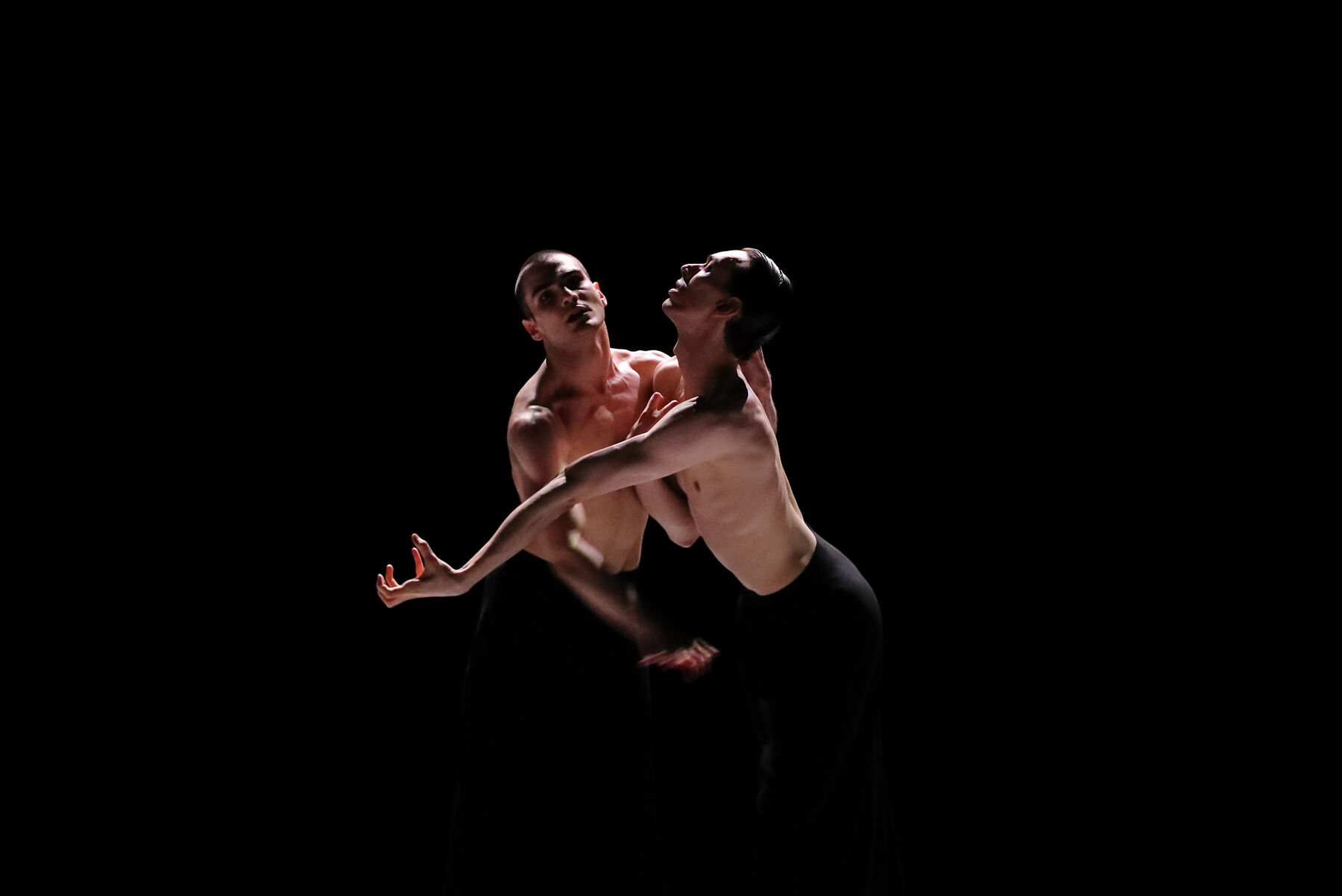Zwei Tänzer auf einer dunklen Bühne.  Sie stehen sich mit verschlungenen Armen gegenüber, die Gesichter voneinander abgewendet. Beide tragen schwarze Hosen und sind oberkörperfrei.