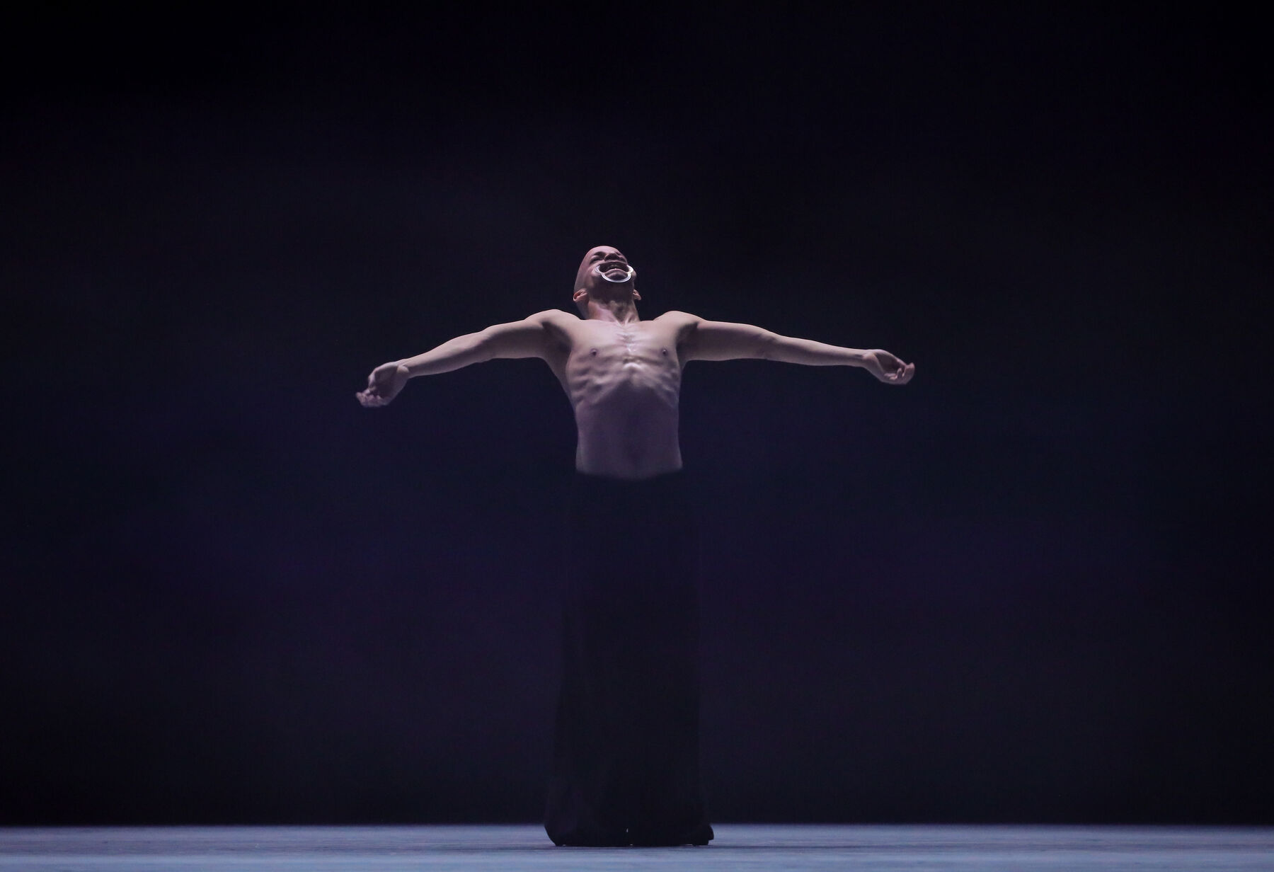 Ein Tänzer steht auf einer dunklen Bühne. Er trägt nur eine schwarze, weite Hose. Er breitet beide Arme aus und lehnt seinen Kopf nach hinten. Sein Mund ist mit einem Wangenspreizer geöffnet. Ein leichtes Licht kommt von oben. 