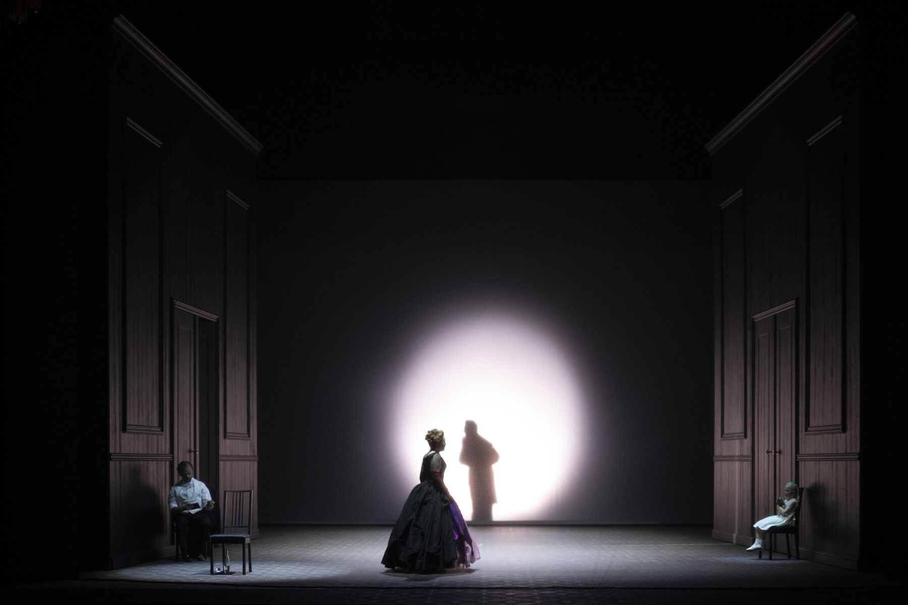 Eine Frau steht in einem dunklen Raum und schaut auf einen Lichtkegel an der Wand, auf dem sich der Schatten eines Mannes abzeichnet.