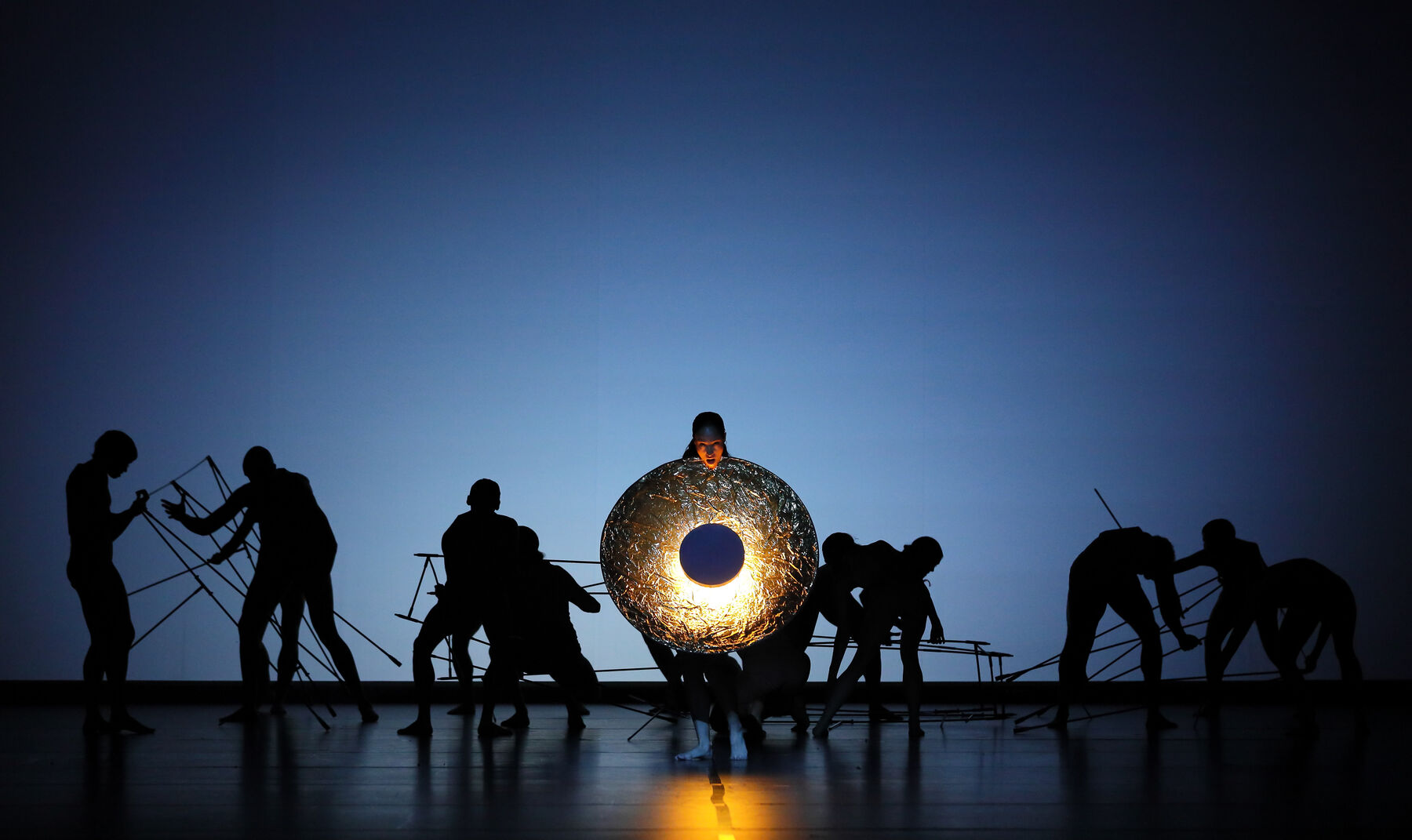 A dancer holds a glowing golden disc. In the background, the silhouettes of other dancers can be seen wielding poles. The background is blue.