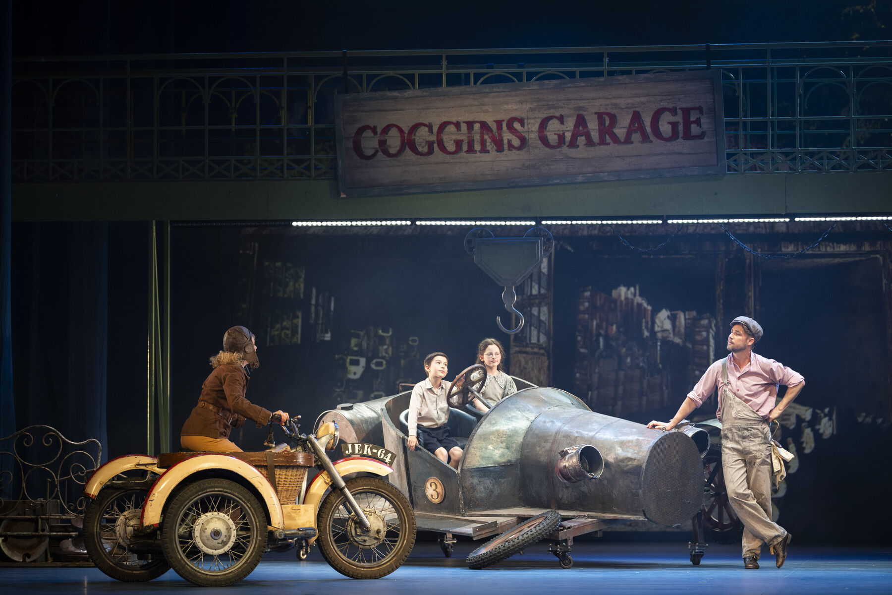 A workshop signposted ‘Coggins Garage’. Two children are sitting in a broken-down old car. A man is leaning casually against the bonnet, watching a woman slowly approaching them on a motorbike with a sidecar. 