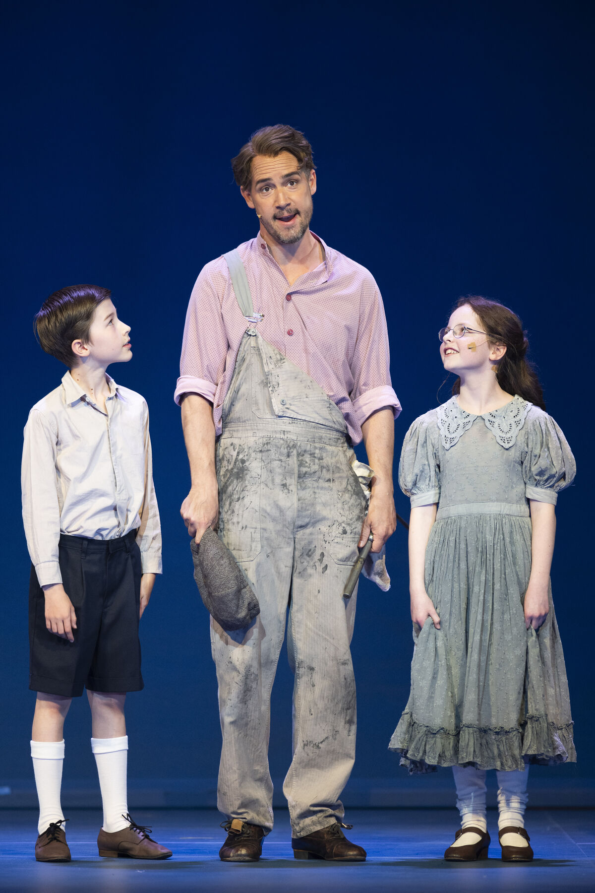 A man in dirty work clothes stands between a boy and a girl who look at him expectantly.