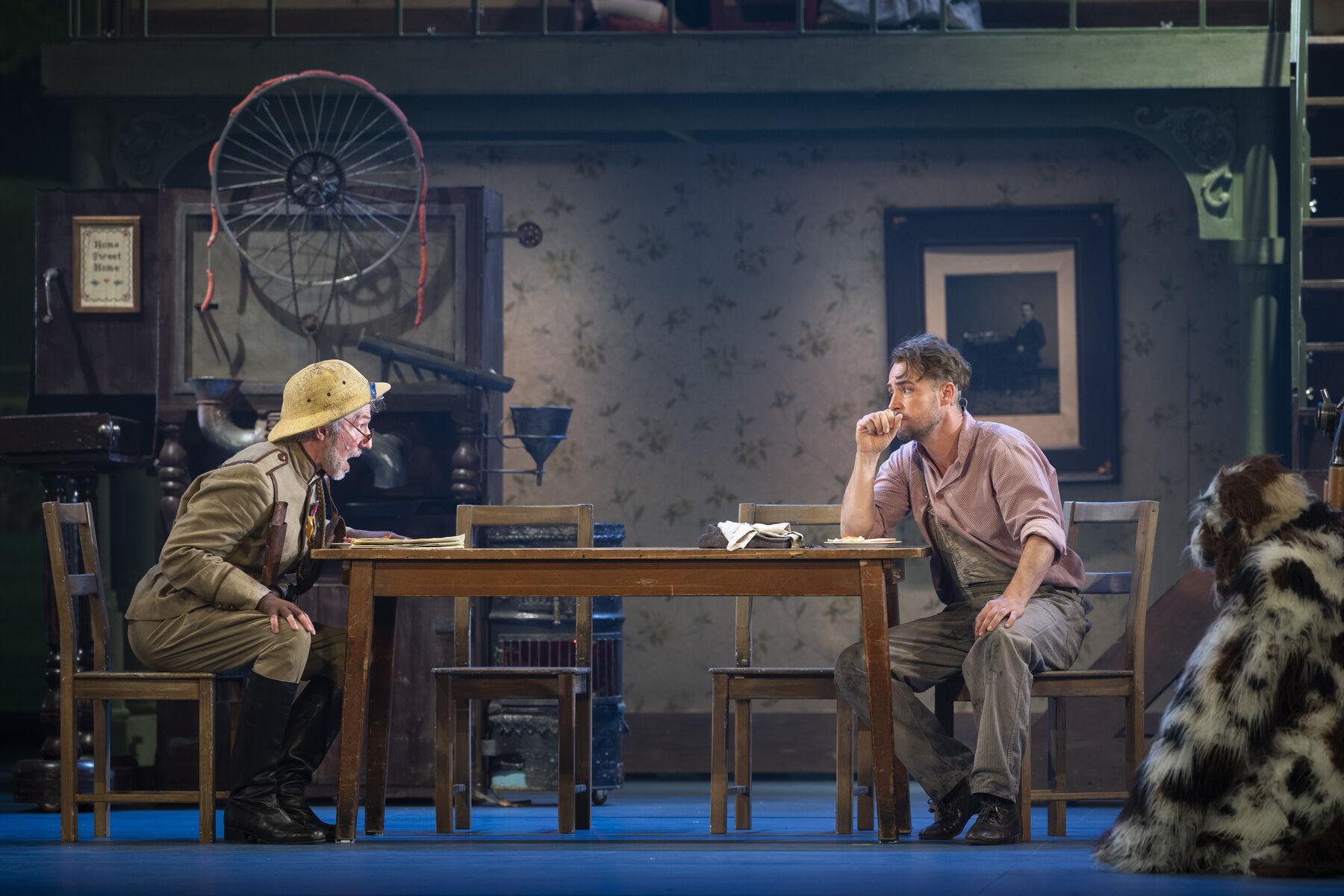 Two men are sitting opposite each other, the younger one looks worried and rests his hand on his chin. The older one leans forward invitingly. The scene takes place in a living room. 