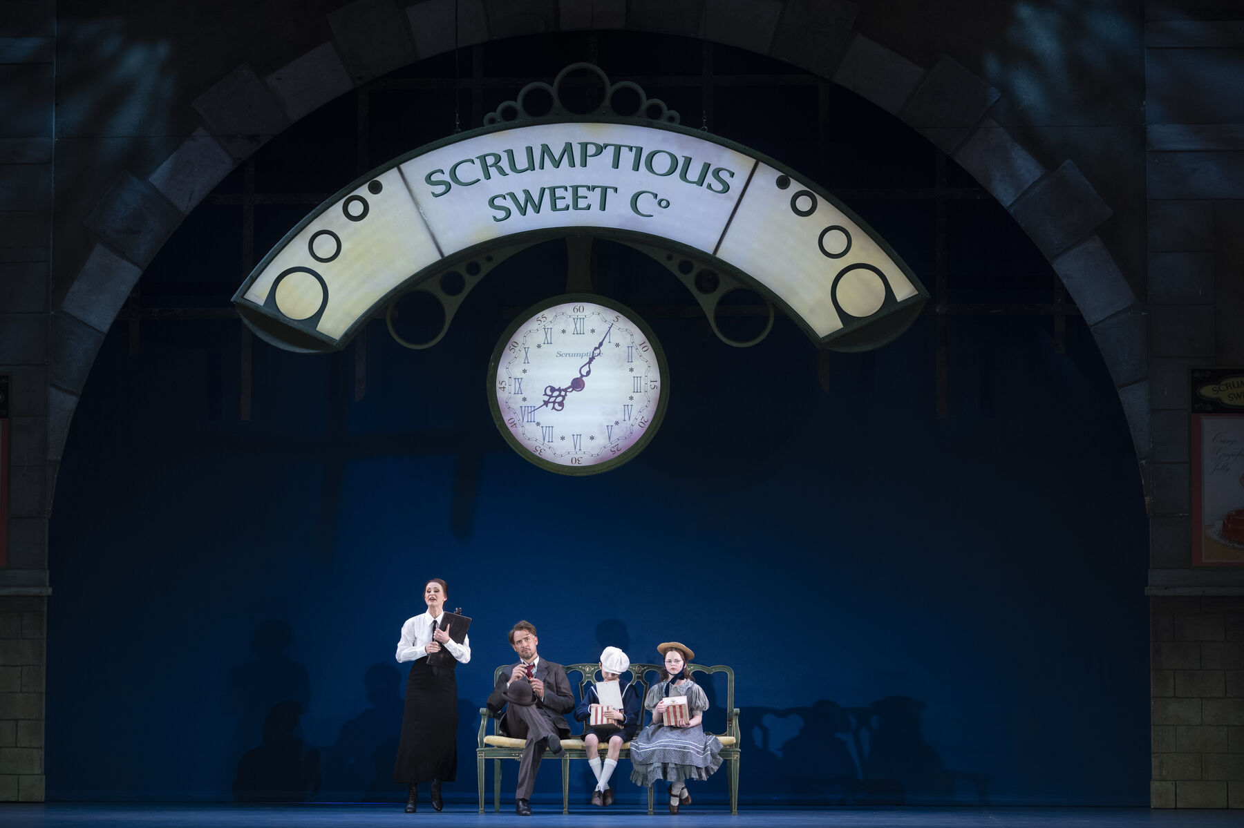 A man and two children are sitting on a bench. Next to them stands a stern-looking woman holding a clipboard. Above them hangs a large clock and a reed with the inscription ‘scrumptious sweet c°’.