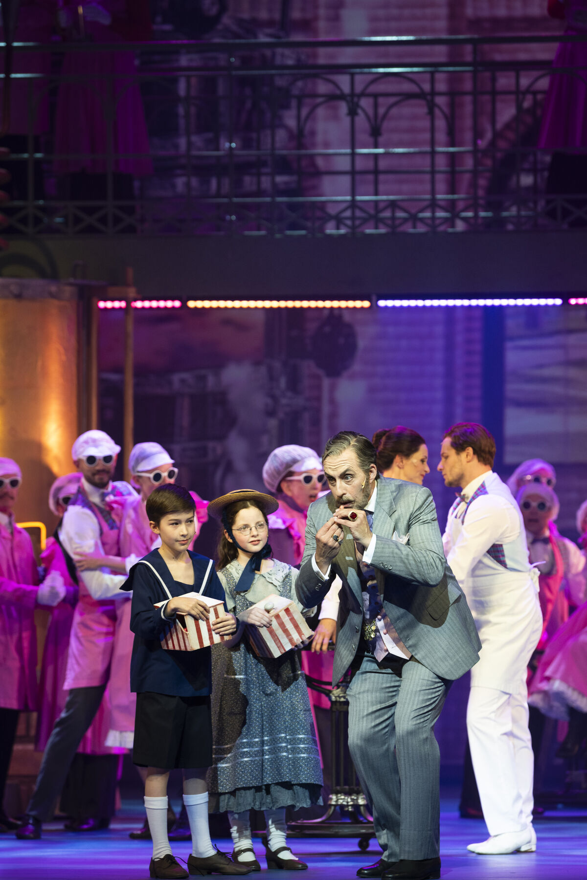 An elderly man blows into a small flute. The two children look at him expectantly. Several factory workers can be seen in the background.