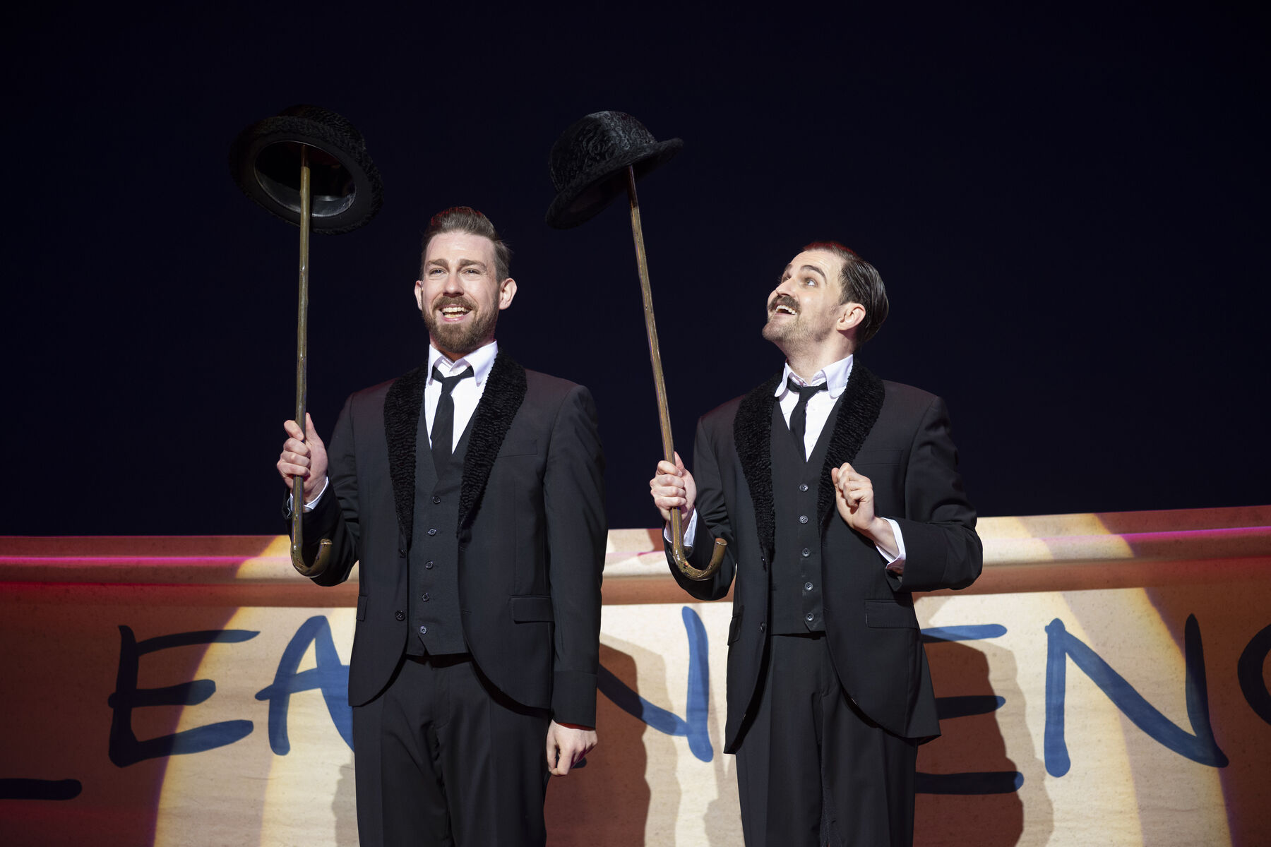 Two men in black tuxedos stand next to each other, balancing bowler hats on walking sticks. 