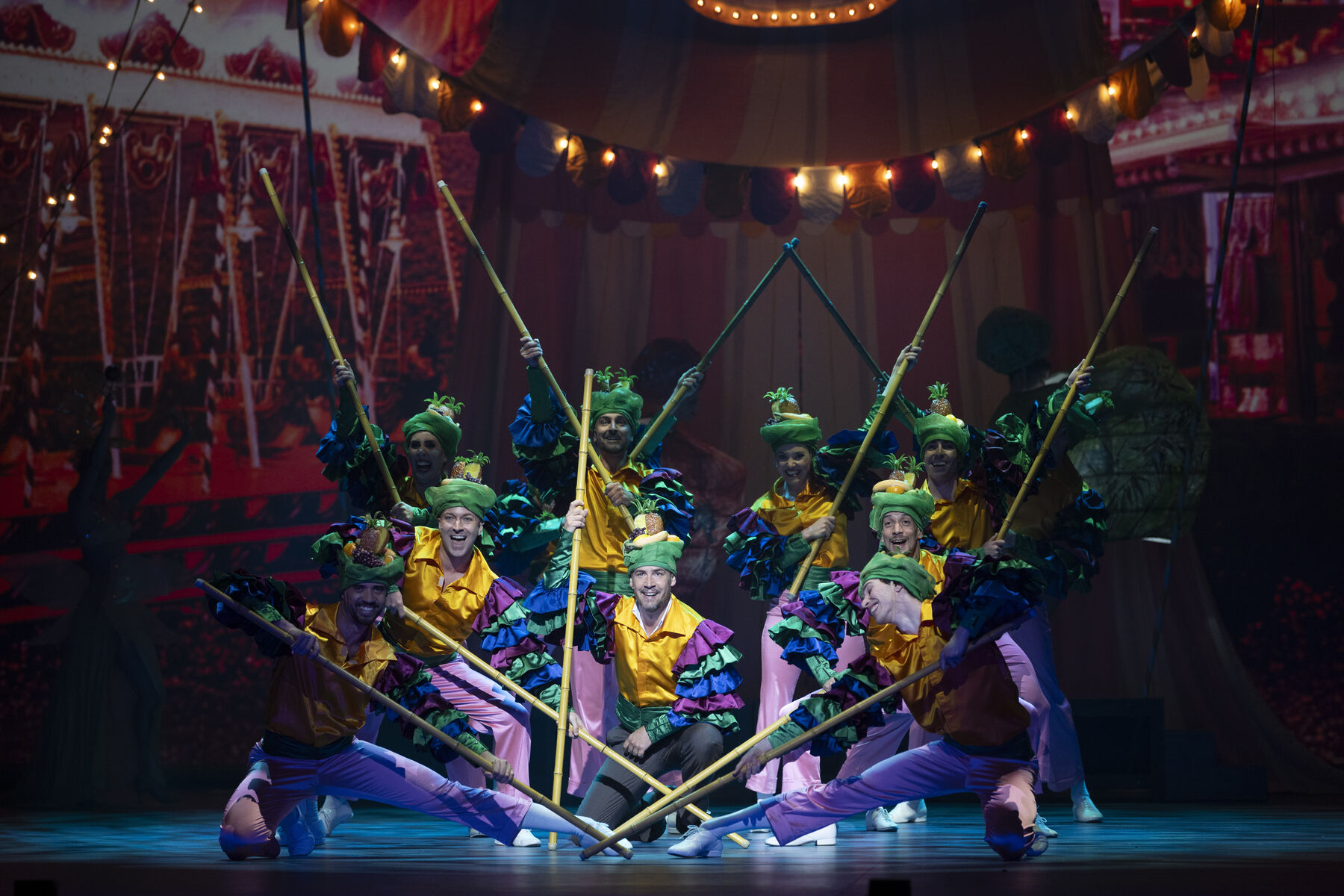 A group of men in colourful costumes pose with large bamboo sticks in a circus. 