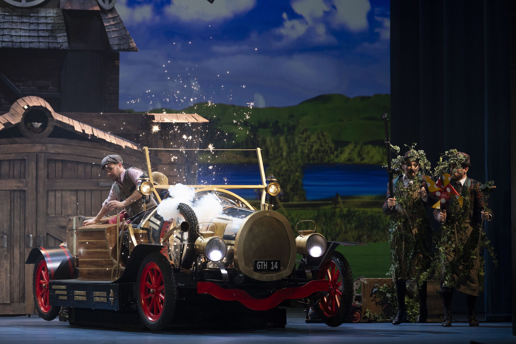 A man is sitting in a car, sparks are flying. Two men in camouflage clothing are standing next to him. A windmill can be seen in the background.