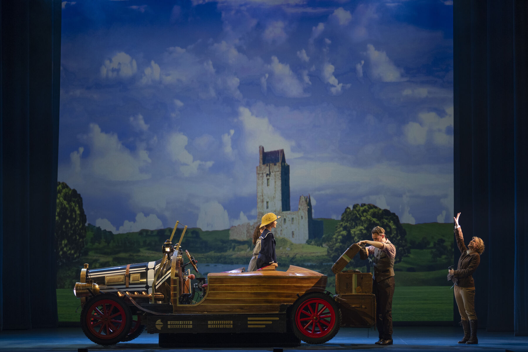 Two children are standing on the back seat of a Caprio, looking back at a man who is closing the boot.  Behind him stands a woman, smiling and waving at the children. In the background, you can see a castle standing in a green landscape.