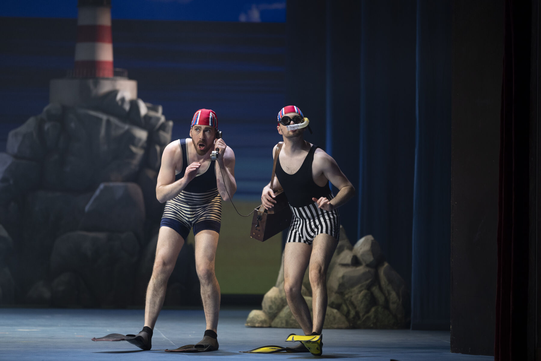 Two men in striped swimsuits with red swimming caps and diving fins can be seen. The man on the right is carrying a suitcase with a portable telephone. The man on the left is holding the receiver in his hand and looking around. In the background are a lighthouse, rocks and the sea.