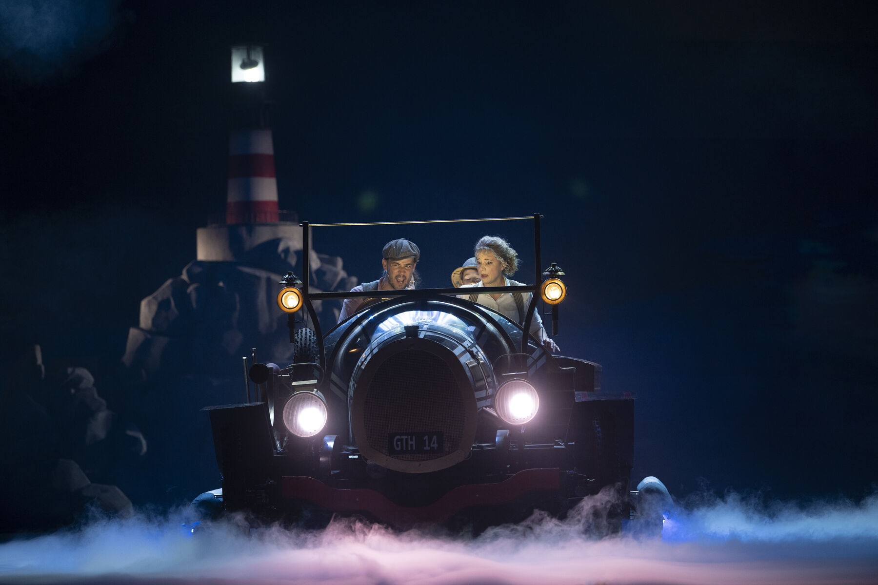 Two adults and two children are sitting in a car. In the background, you can see a lighthouse at night. Fog covers the ground. The man at the wheel seems upset, and the woman next to him looks very worried.