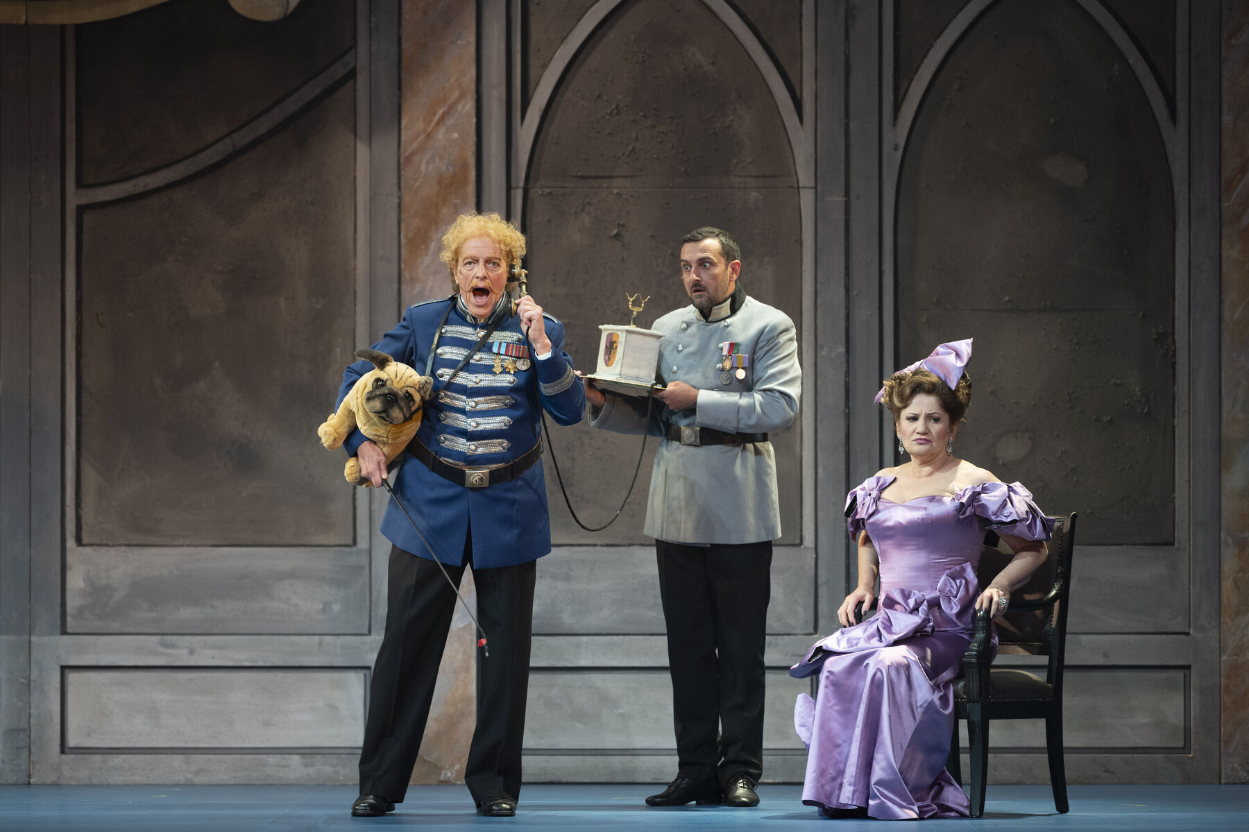 A man in a general's uniform is shouting into a telephone. He is holding a stuffed toy of an English bulldog under his arm. A servant is holding the telephone for him and looks frightened. A middle-aged woman in a lilac evening gown sits unhappily on a chair and averts her gaze from the shouting man.