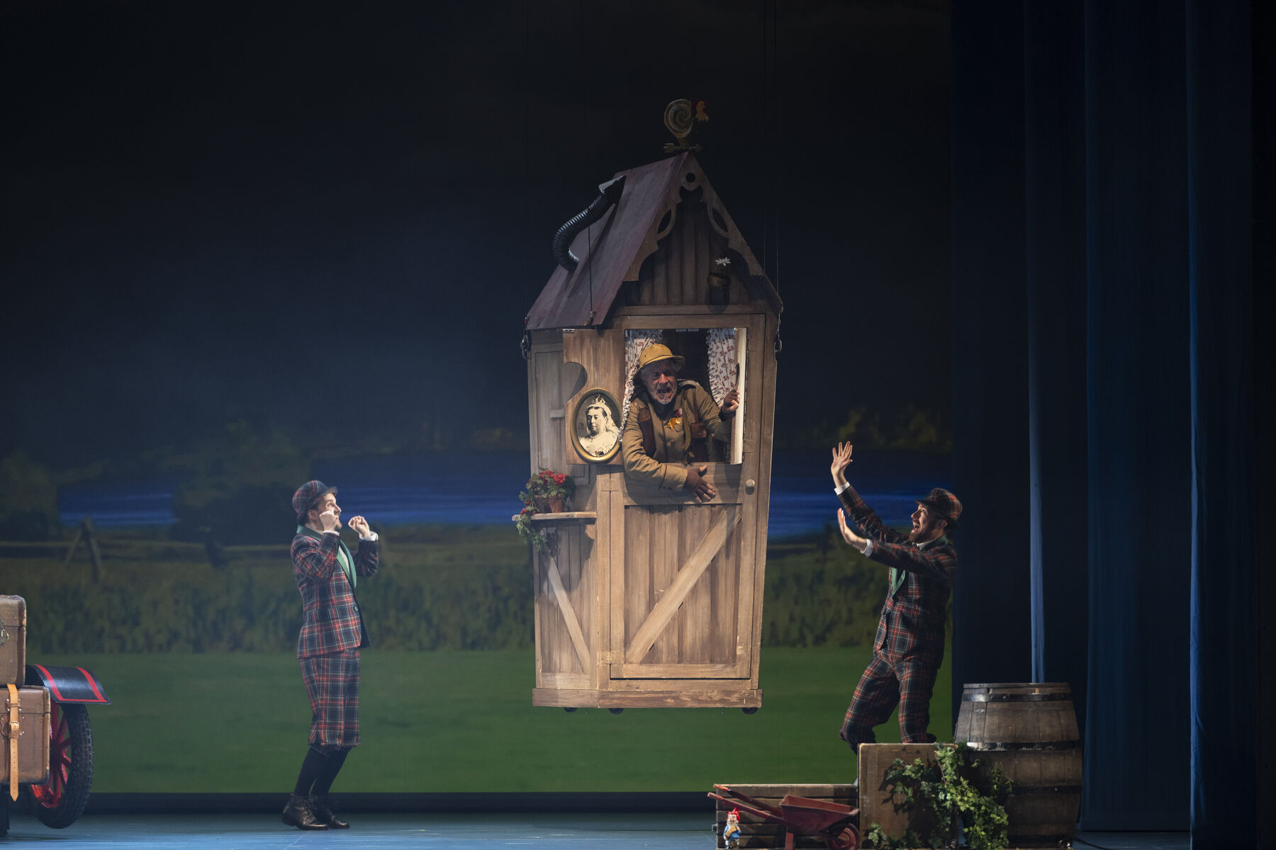 A wooden toilet block hovers above the ground. A man in a ranger uniform looks out of the open window in alarm. To the left and right of the building, two men in tartan suits stand looking pleased. In the background is a green landscape and a small lake.