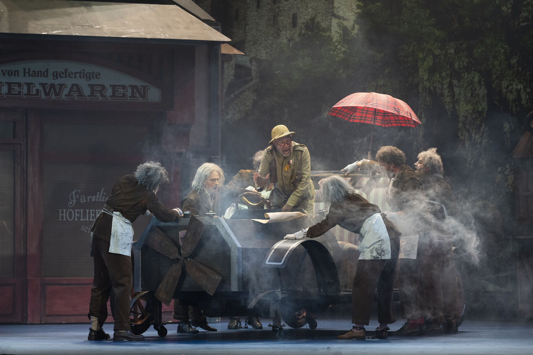 An elderly man in a ranger uniform stands in front of a plan in the body of an old car. He raises his open hand in agitation. Around the car, many elderly men in monotonous work clothes stand and touch the bodywork. In the background is a toy shop.