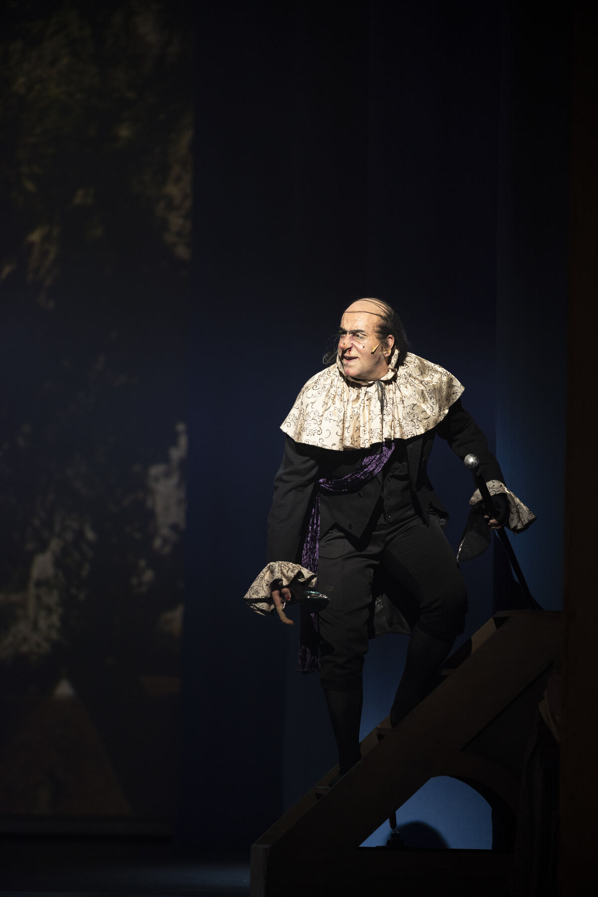 The picture shows a man in a black tailcoat. He has a large white collar and white sleeves protruding from it. He is balding and has black hair that reaches approximately to his chin. His nose is long and pointed, and he has several moles on his face. He looks angry. The stage is dark.