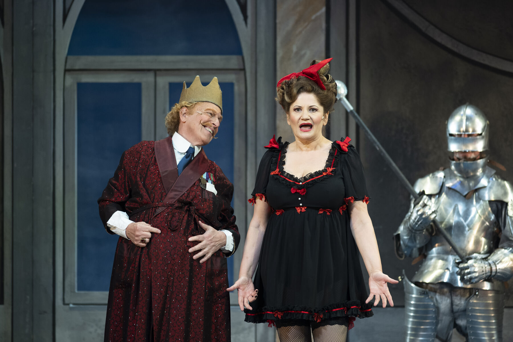 A man in uniform is wearing a red dressing gown and looking at his wife with a grin. He has a cardboard crown on his head. The woman standing next to him is wearing a short black Lolita-style dress with red bow decorations and fishnet stockings. She looks slightly annoyed and is gesturing. Behind the couple stands a knight in silver armour. There is a large window in the background.