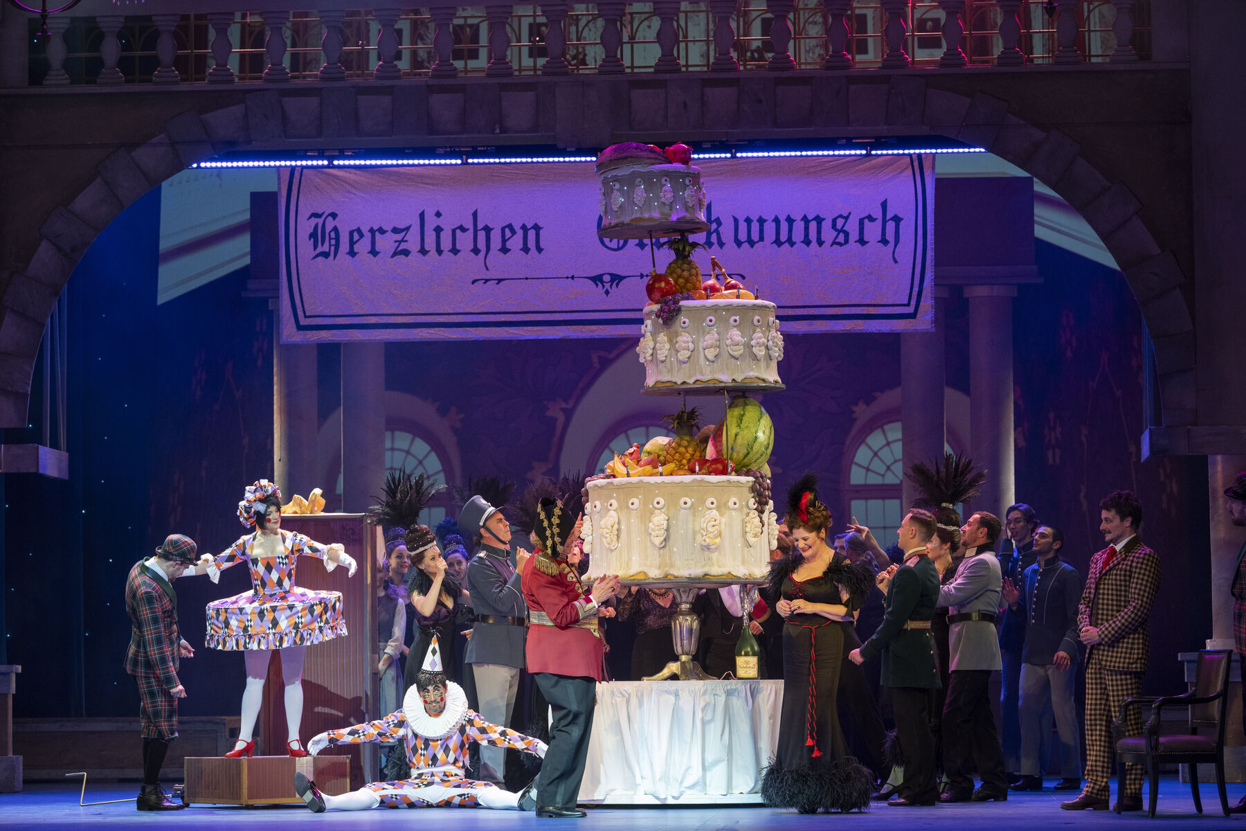 A celebration with lots of people. A large banner hangs from the ceiling with the words ‘Congratulations’. In the middle of the room is a huge multi-tiered cake.