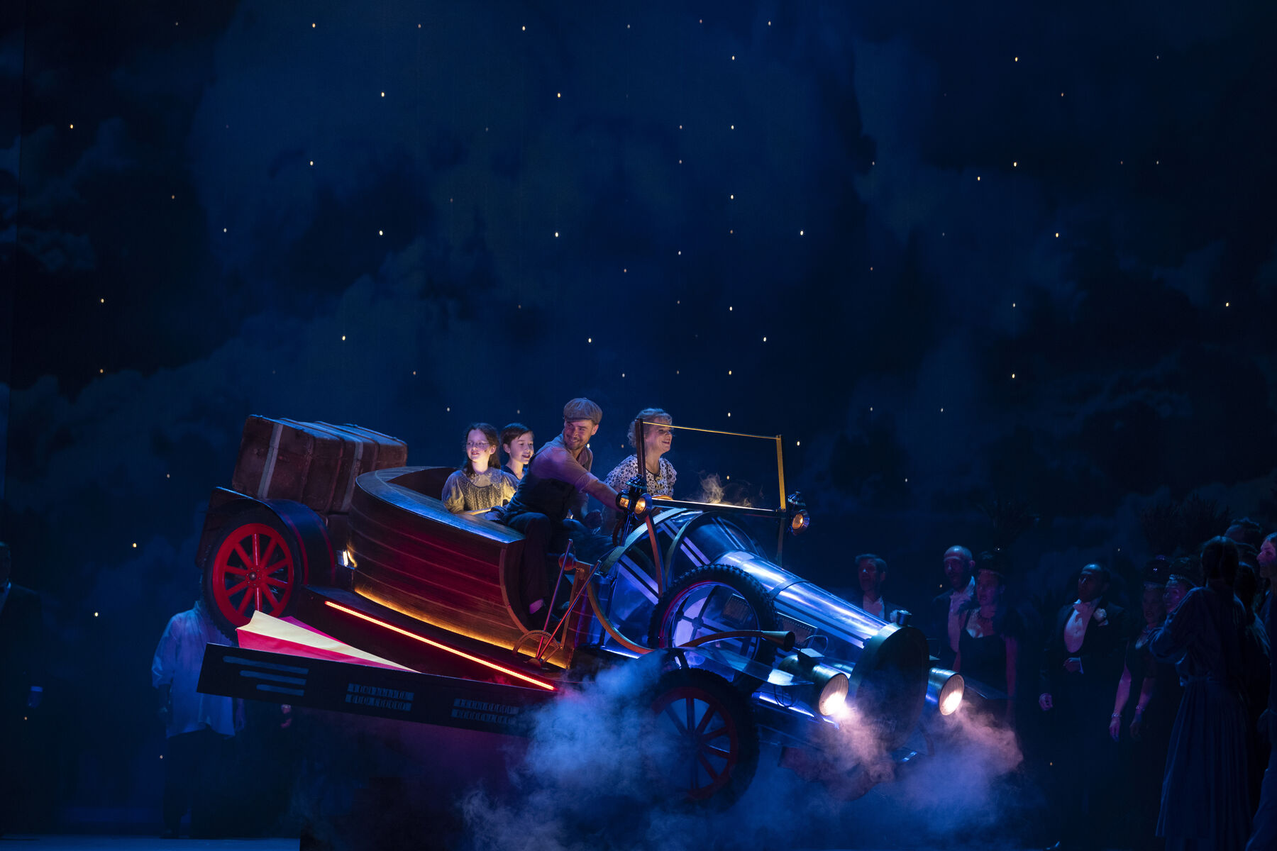 Two adults and two children are sitting happily in a flying car. A group of people are standing together on the ground, looking at the car in surprise. The scene takes place at night.