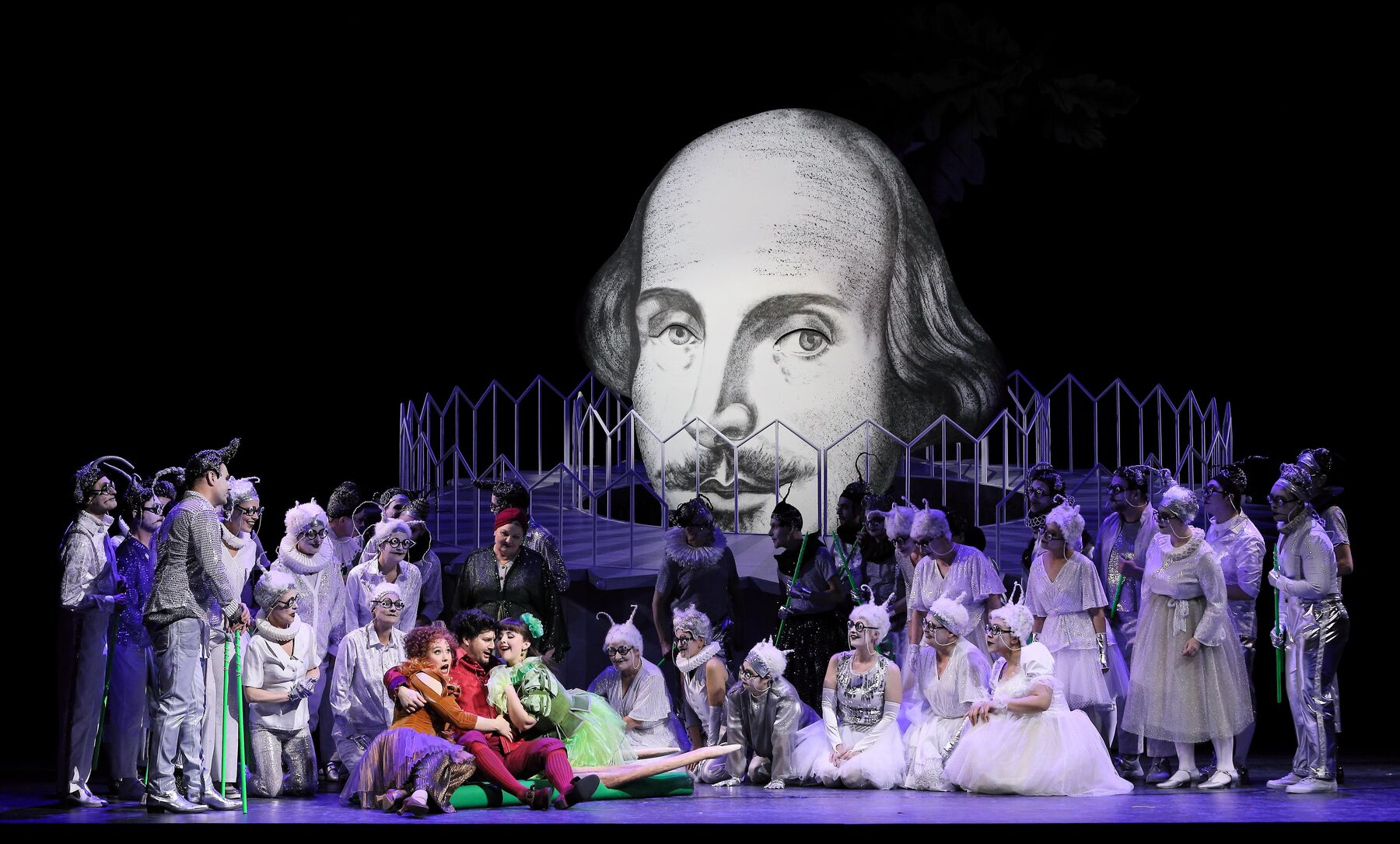 Frau Fluth, Sir John Falstaff und Frau Reich sitzen auf der Bühne. um sie herum, in weiß, ist der Chor verteilt. Im Hintergrund sieht man das Gesicht von Shakespeare.