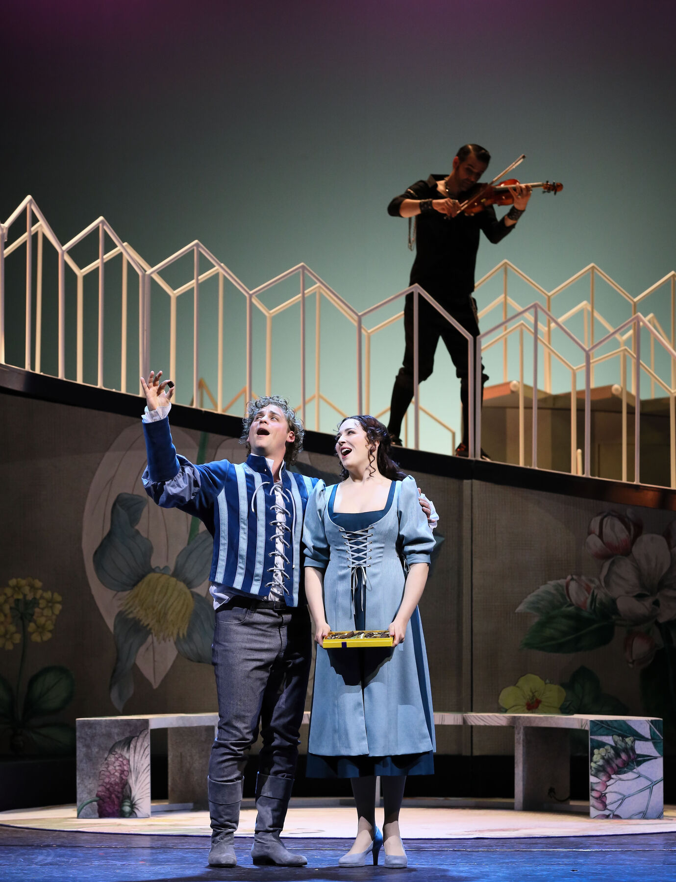 Fenton und Anna Reich stehen in blauer Kleidung auf der Bühne und singen. Hinter den beiden auf einem Übergang ist ein Kellner mit Geige in schwarz zu sehen.