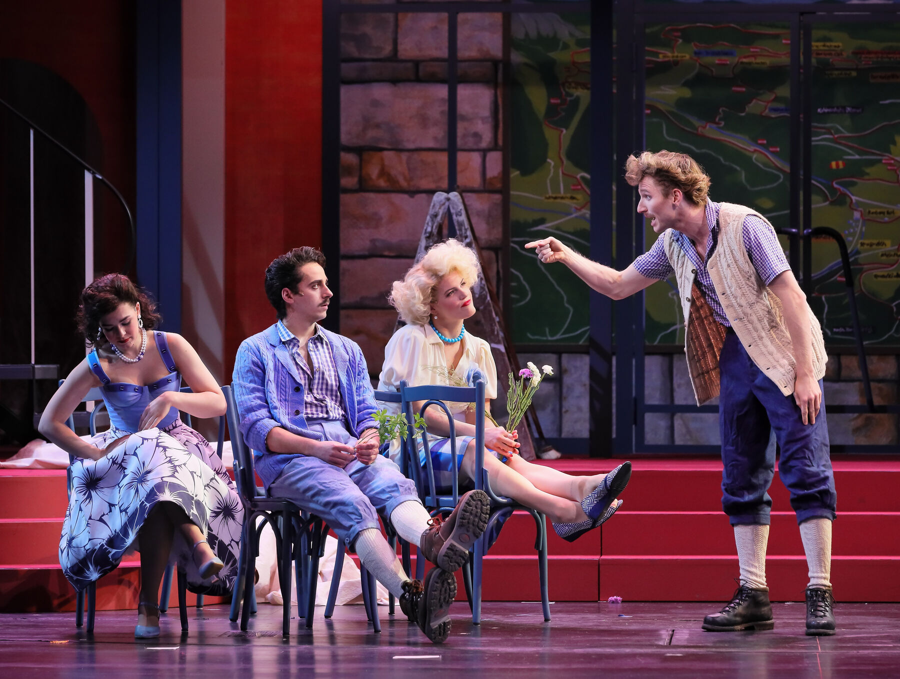 Botho sits on a chair between Pauline and Regina. He is holding a bouquet of woodruff in his hand. Erich stands in front of them and points his index finger accusingly at Botho.