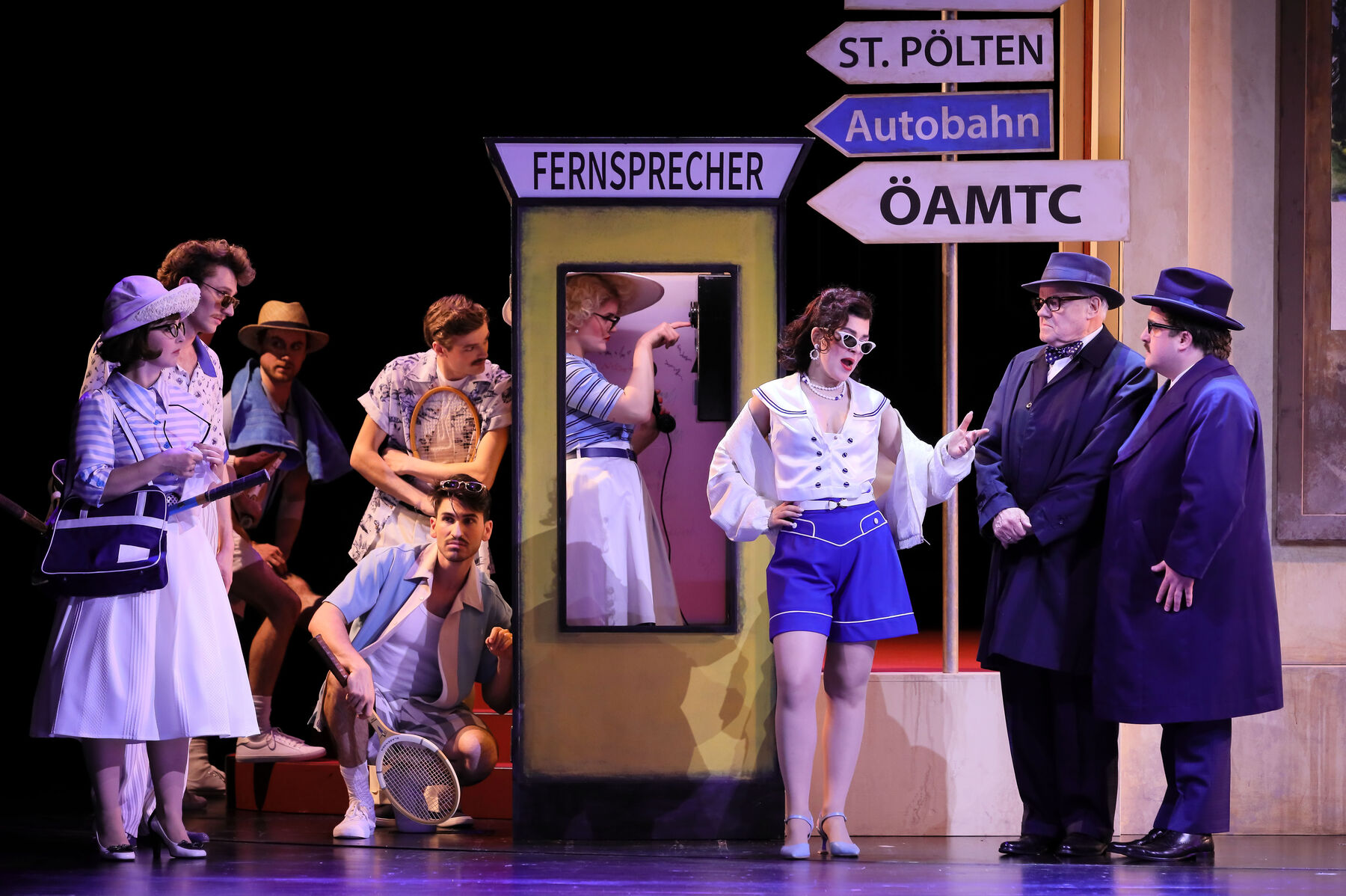 A street scene. Several young people on the left are looking at Pauline, who is talking to two gentlemen on the right of the picture. The gentlemen are Councillor Danner and Christof Heffele. Pauline is gesturing expansively while the gentlemen stand very stiffly and eye her critically. In the background, a young woman named Regina is standing in a yellow telephone box marked “Fernsprecher” (telephone) and dialling a telephone number.