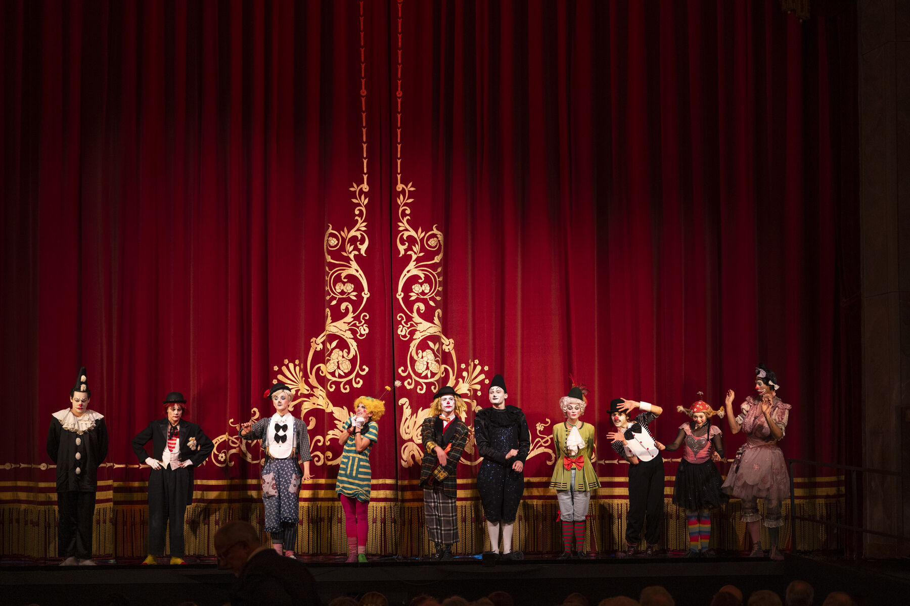 Colourful clowns stand in front of a closed theatre curtain.