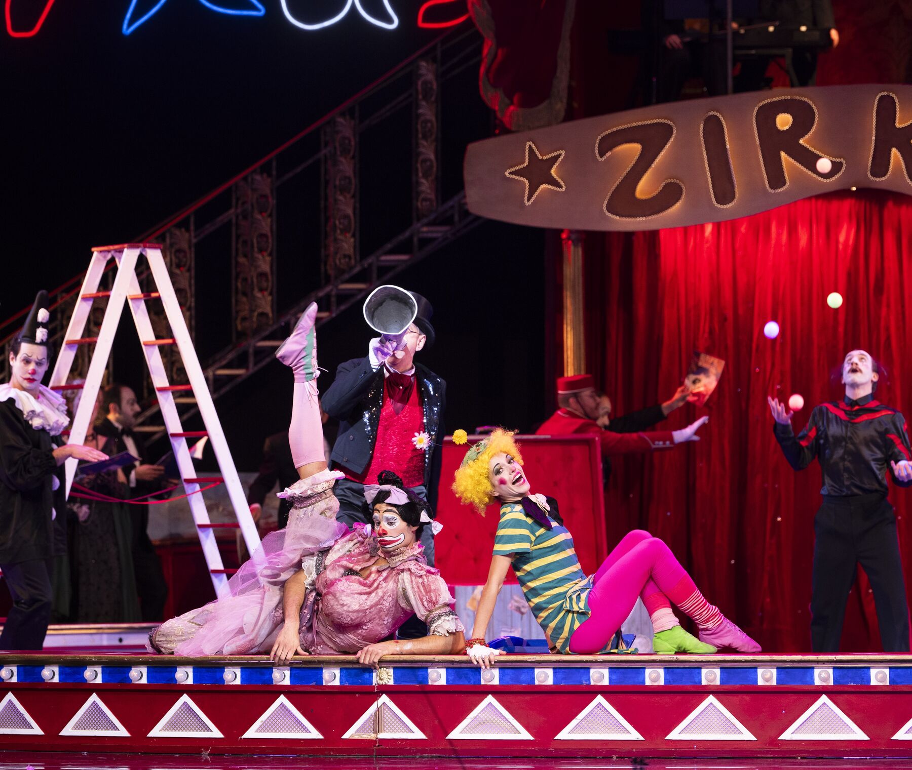 Two clowns sit at the edge of a circus ring and pose in front of a man with a megaphone. 