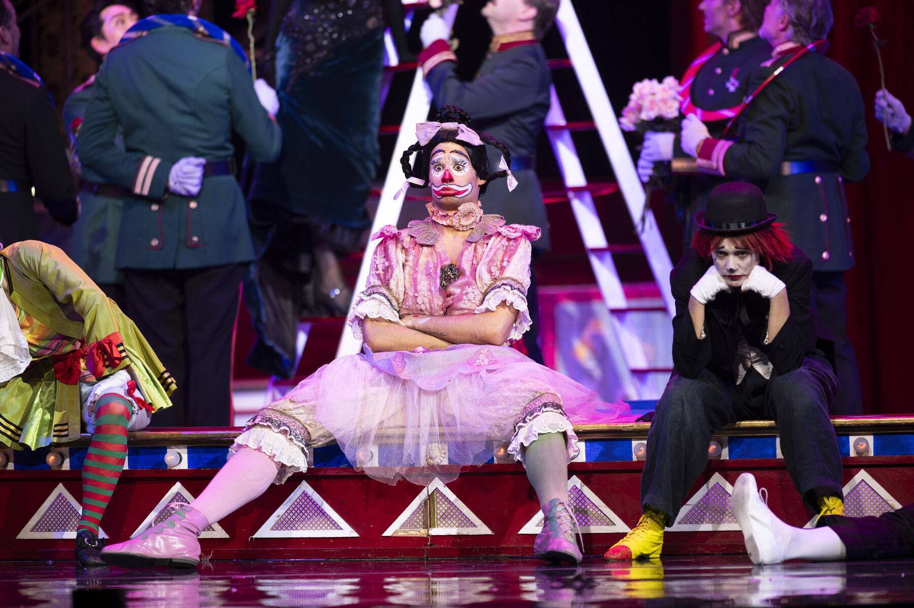 A sulking clown in a pink dress sits with his legs apart and his arms crossed in a circus ring. Next to him sits another clown, sadly lowering his head.