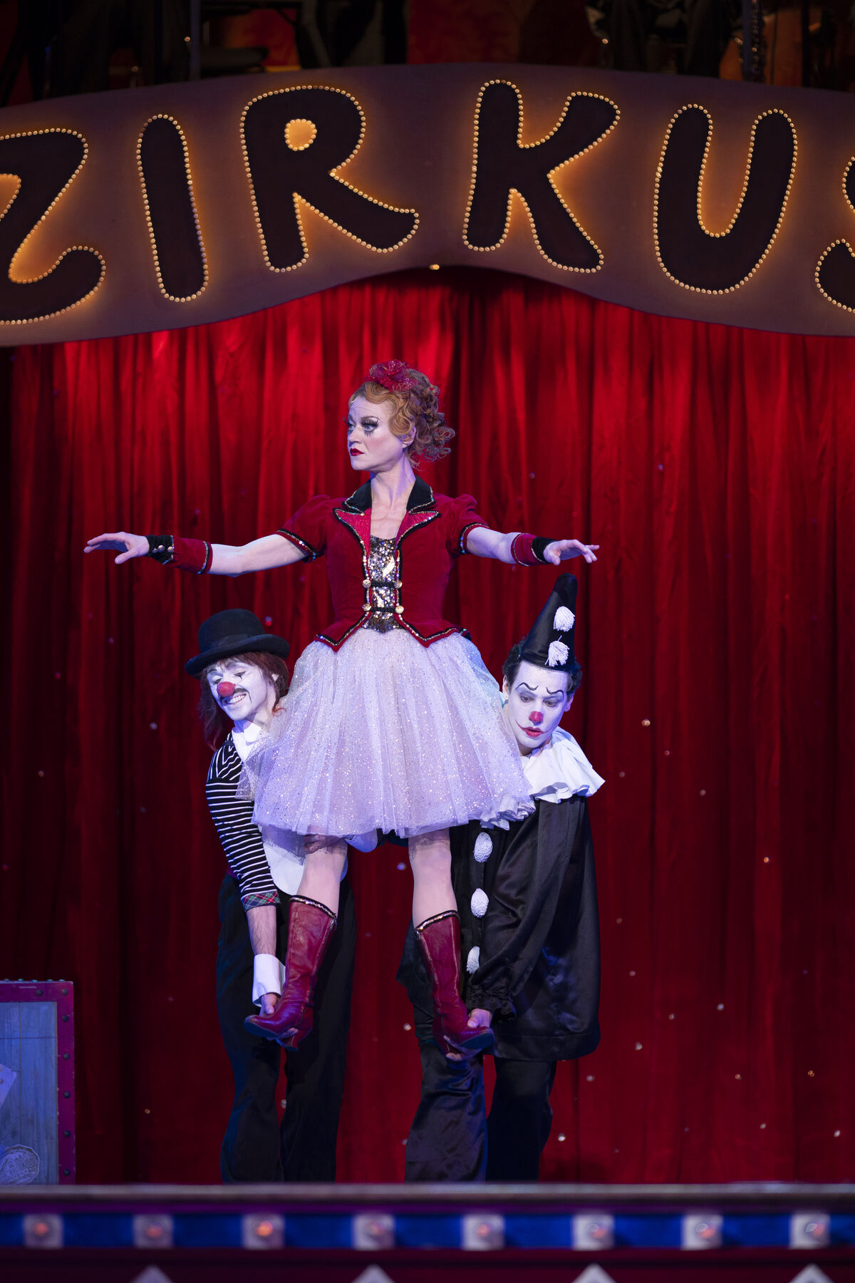 A woman in a red and white dress is carried like a doll by two clowns dressed in black.