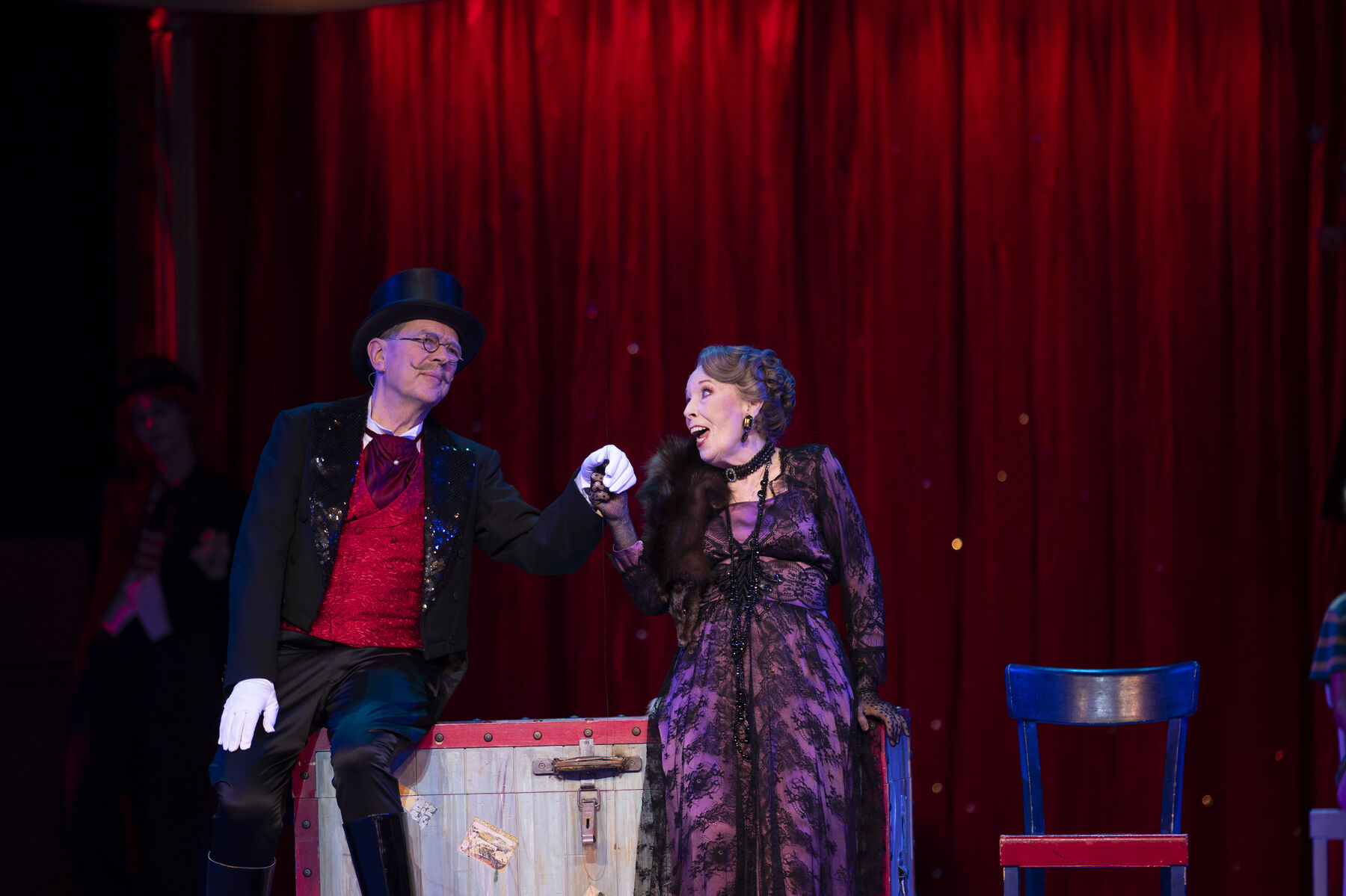 A man wearing a red and black suit and top hat is sitting on a box. Next to him is a woman in a purple dress with black lace. They look at each other and hold hands.