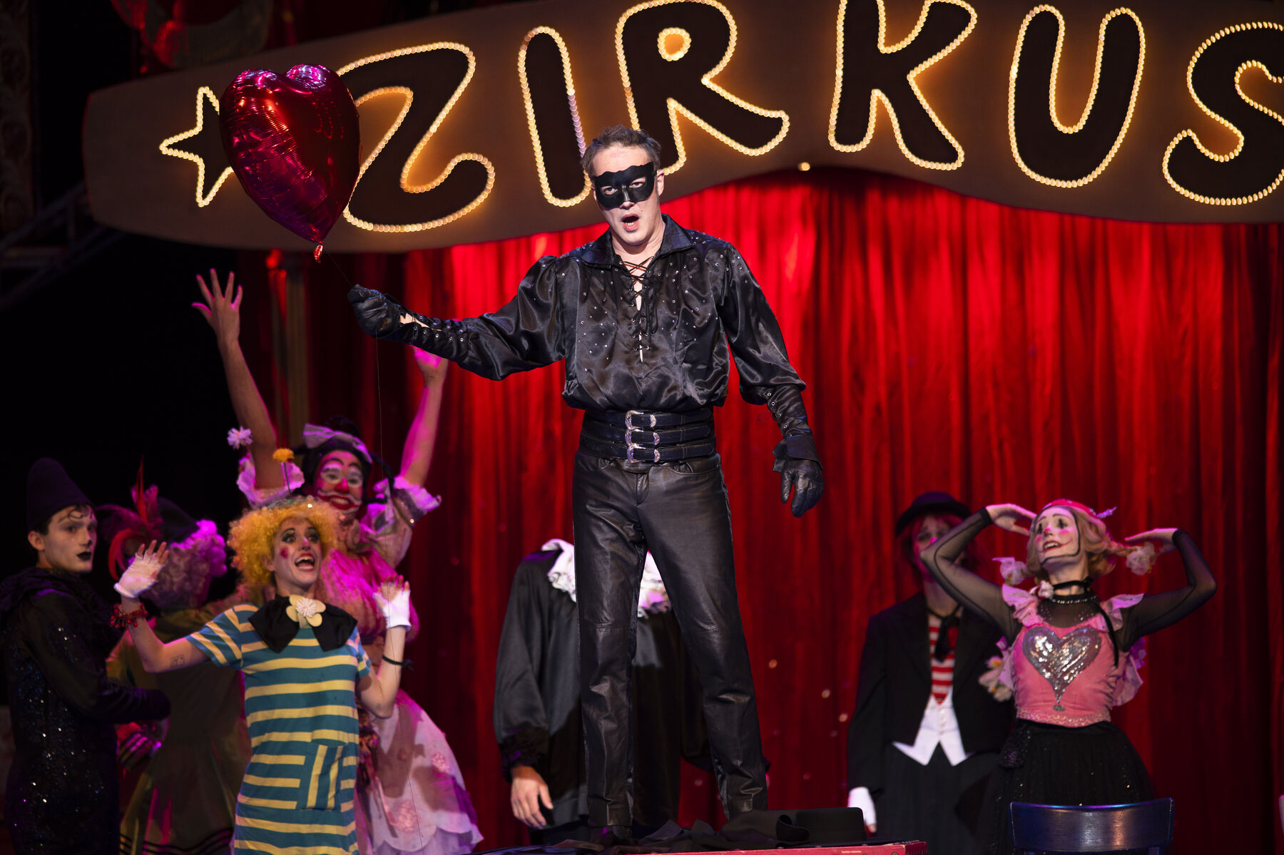 A man in a festive black outfit, wearing a black mask and holding a red heart-shaped balloon, stands on a box. Behind him are cheerful clowns.