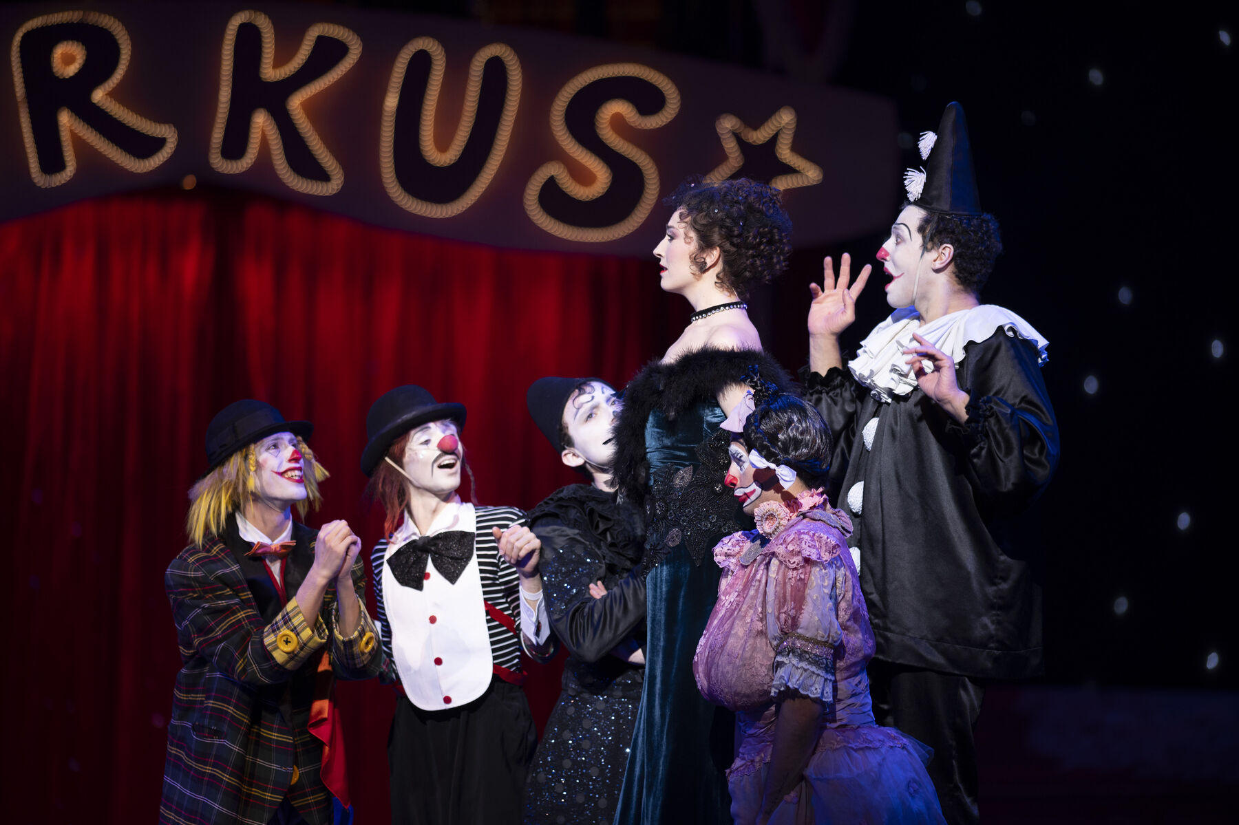 A woman wearing a black and green evening gown stands proudly between five clowns, who look at her joyfully.