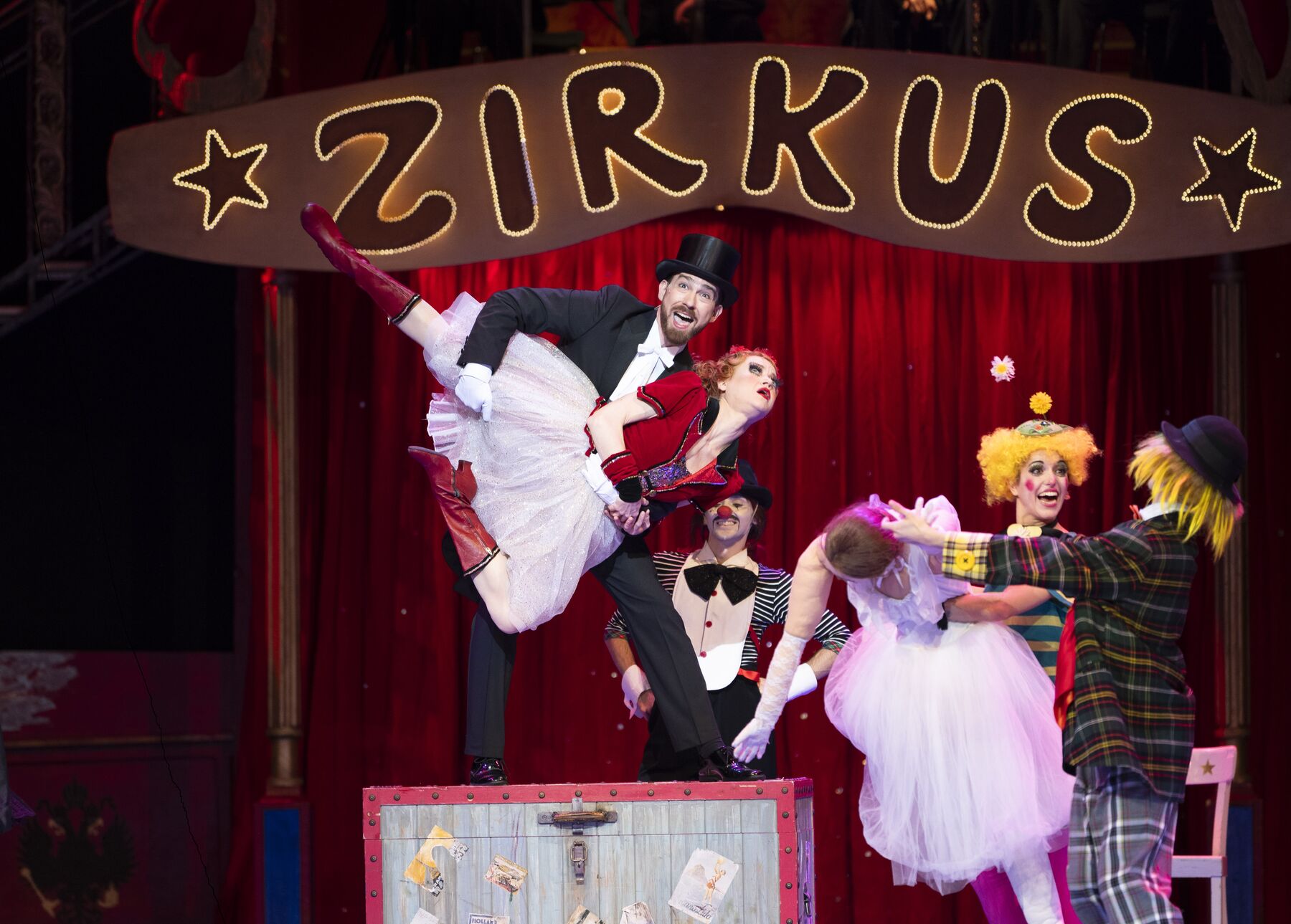 A man in a black suit and black top hat is lifting a woman in a red and white dress. They are standing on a box, surrounded by three clowns.