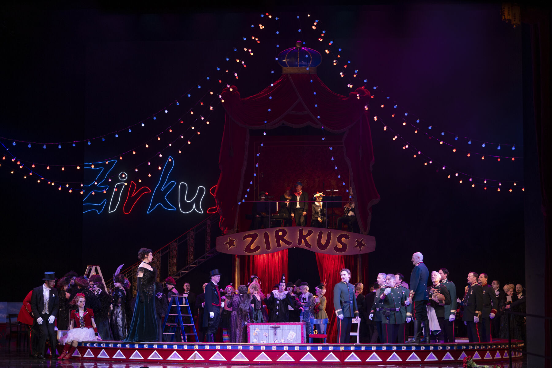 Many different people are standing in a circus ring illuminated by fairy lights. A little further ahead, a woman and a man are standing looking at each other.