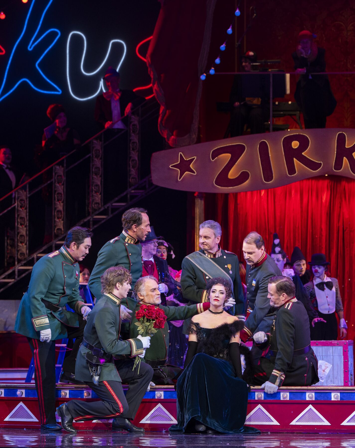 A woman in a black and green evening gown sits sadly in a circus ring. Around her are several concerned men in green uniforms. 