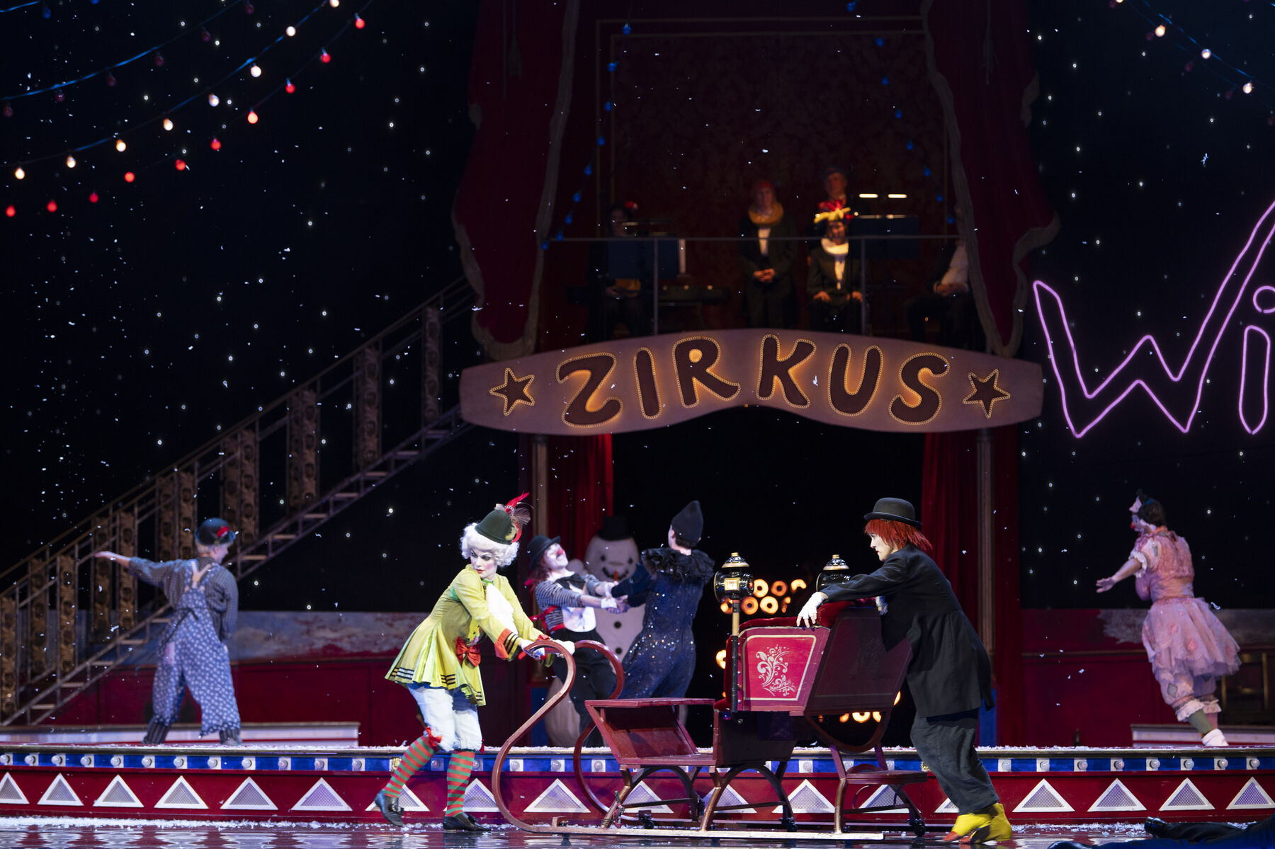 Two clowns push a large sled in front of a circus ring. In the background, more clowns dance in the snow.