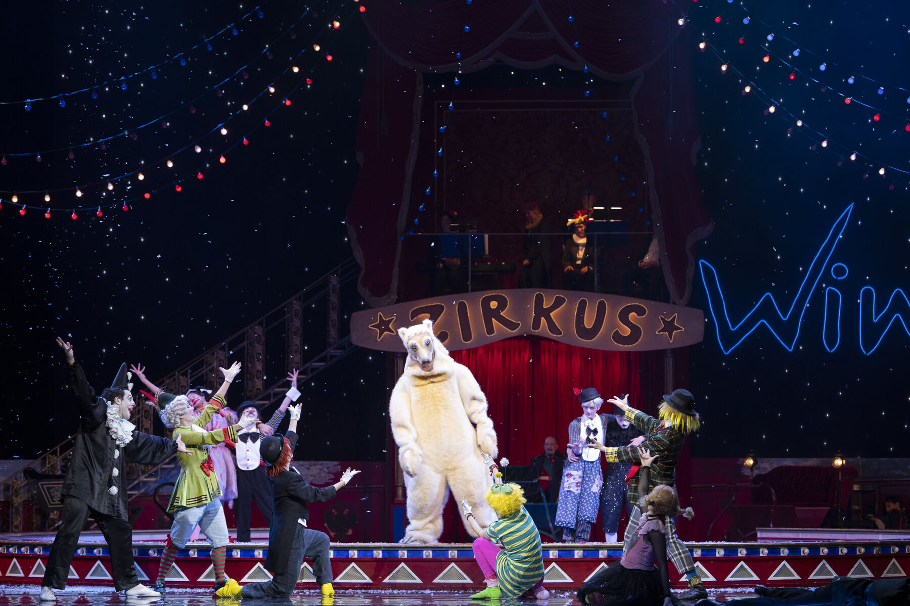 A polar bear stands upright on its hind legs at the edge of a circus ring. Clowns stand around it, pointing at it.