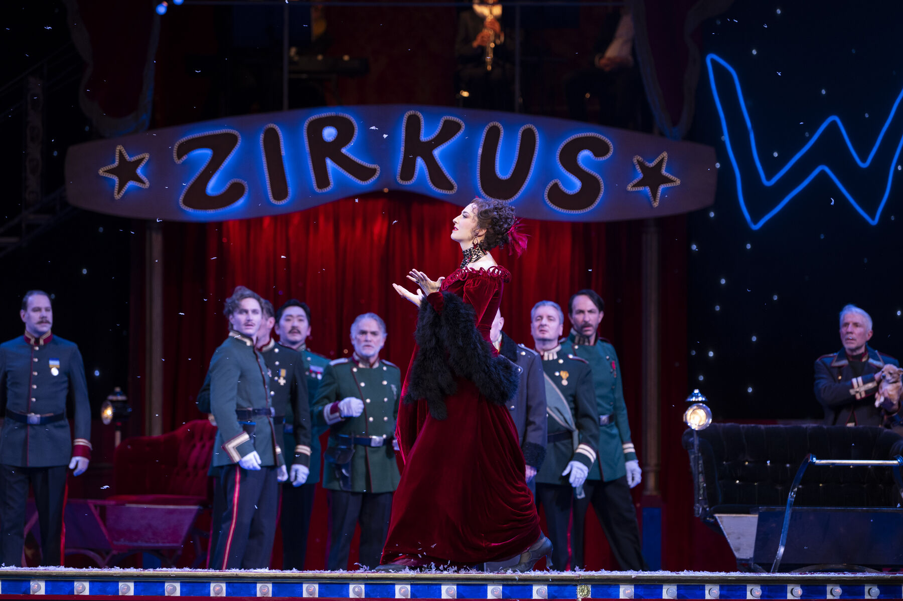 A woman in a red evening gown struts along a circus ring. In the background, men in green uniforms stand watching her.